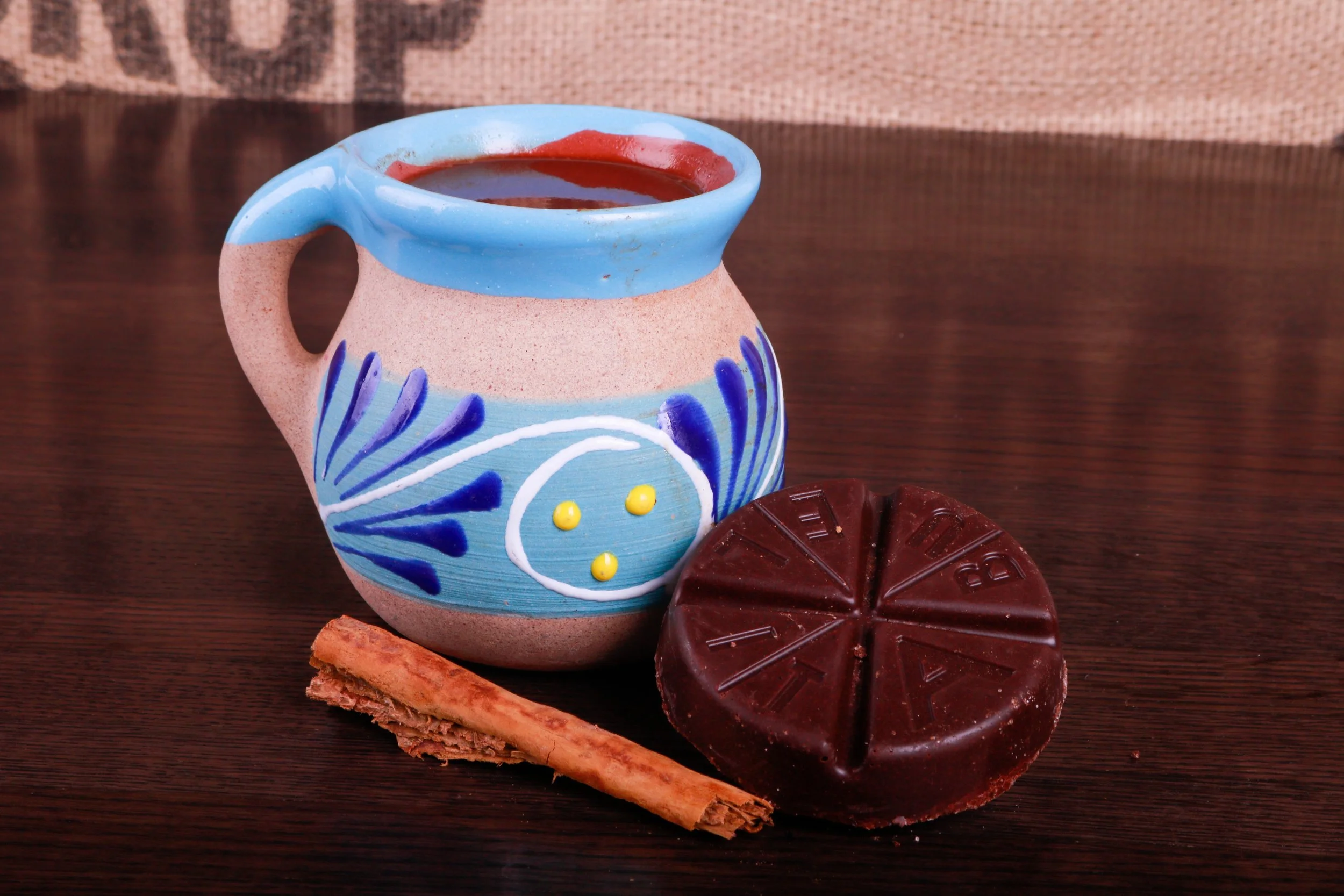 Decorative blue ceramic mug with hot chocolate, a dark chocolate bar, and cinnamon sticks on a wooden table.