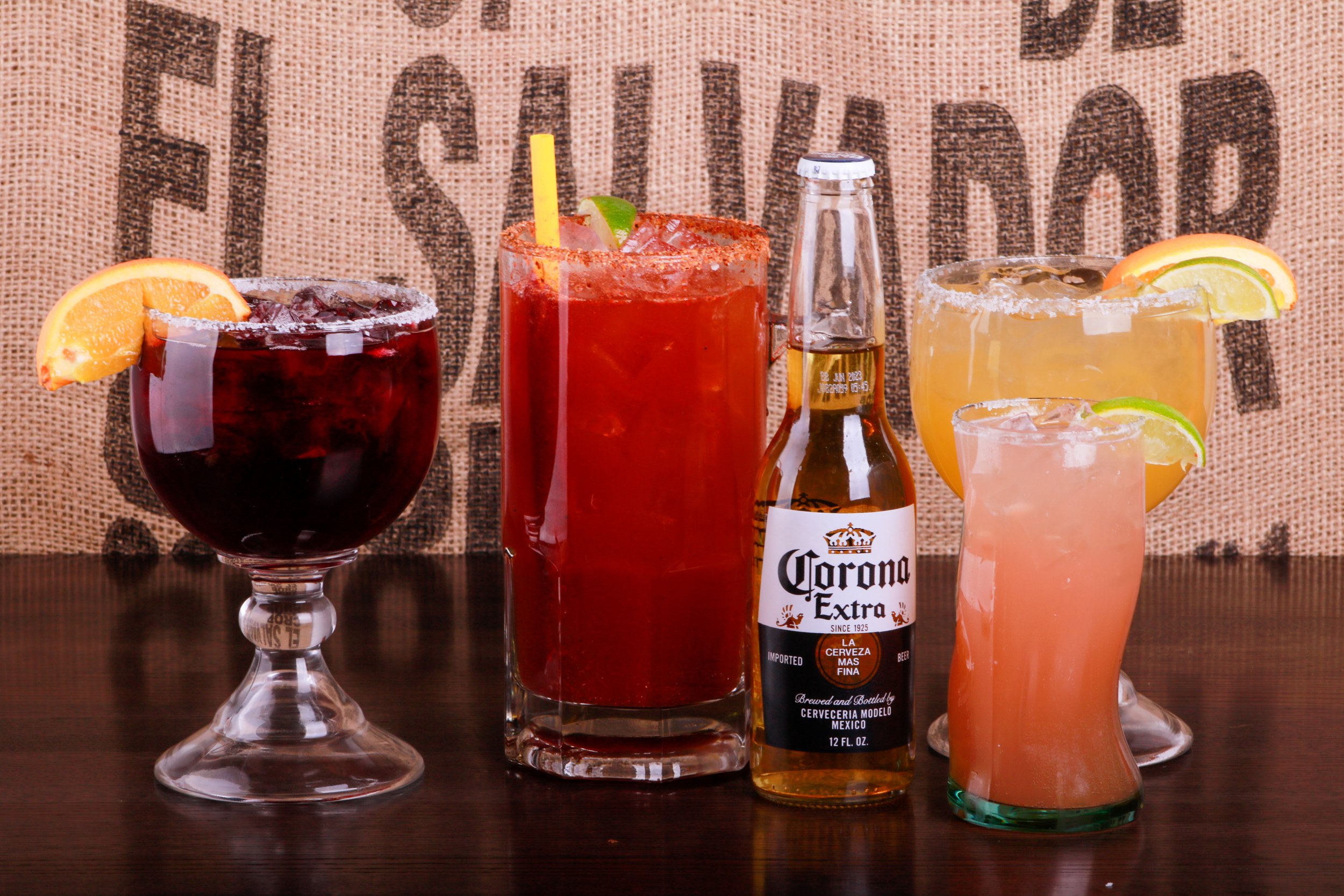 Assorted colorful cocktails with citrus garnishes, a lime-topped red drink, and a Corona Extra beer bottle on a bar.