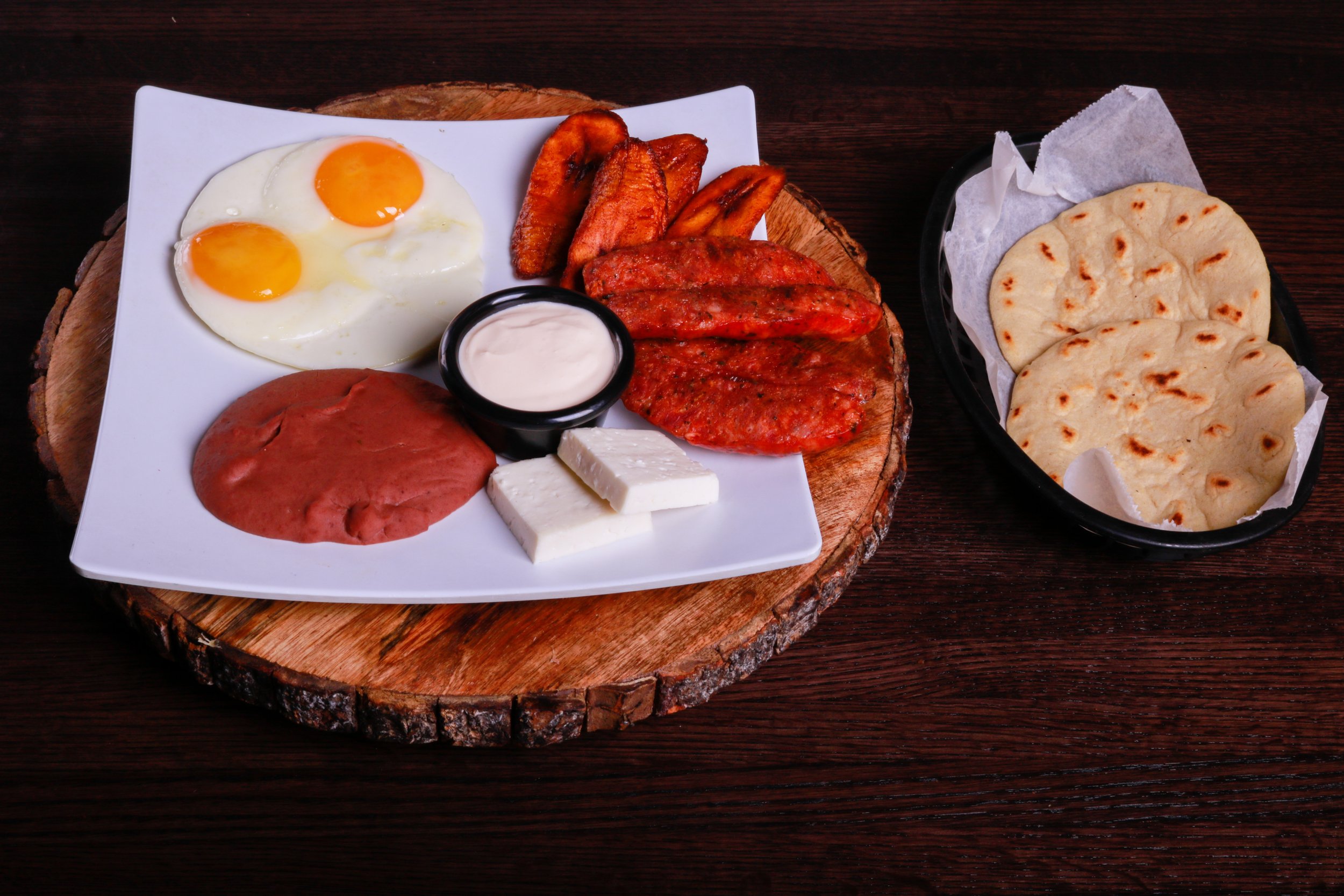 Traditional breakfast platter with fried eggs, grilled sausage, roasted plantains, beans, white cheese, cream sauce, and tortillas