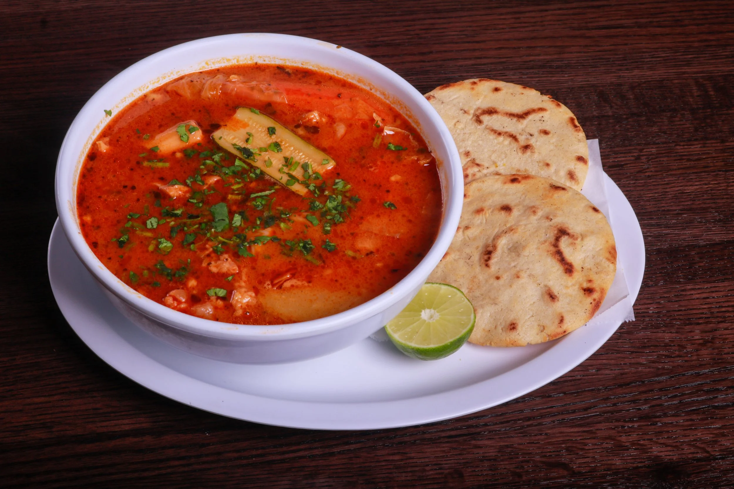 Bowl of spicy red Mexican-style soup garnished with cilantro, served with grilled tortillas and a lime wedge