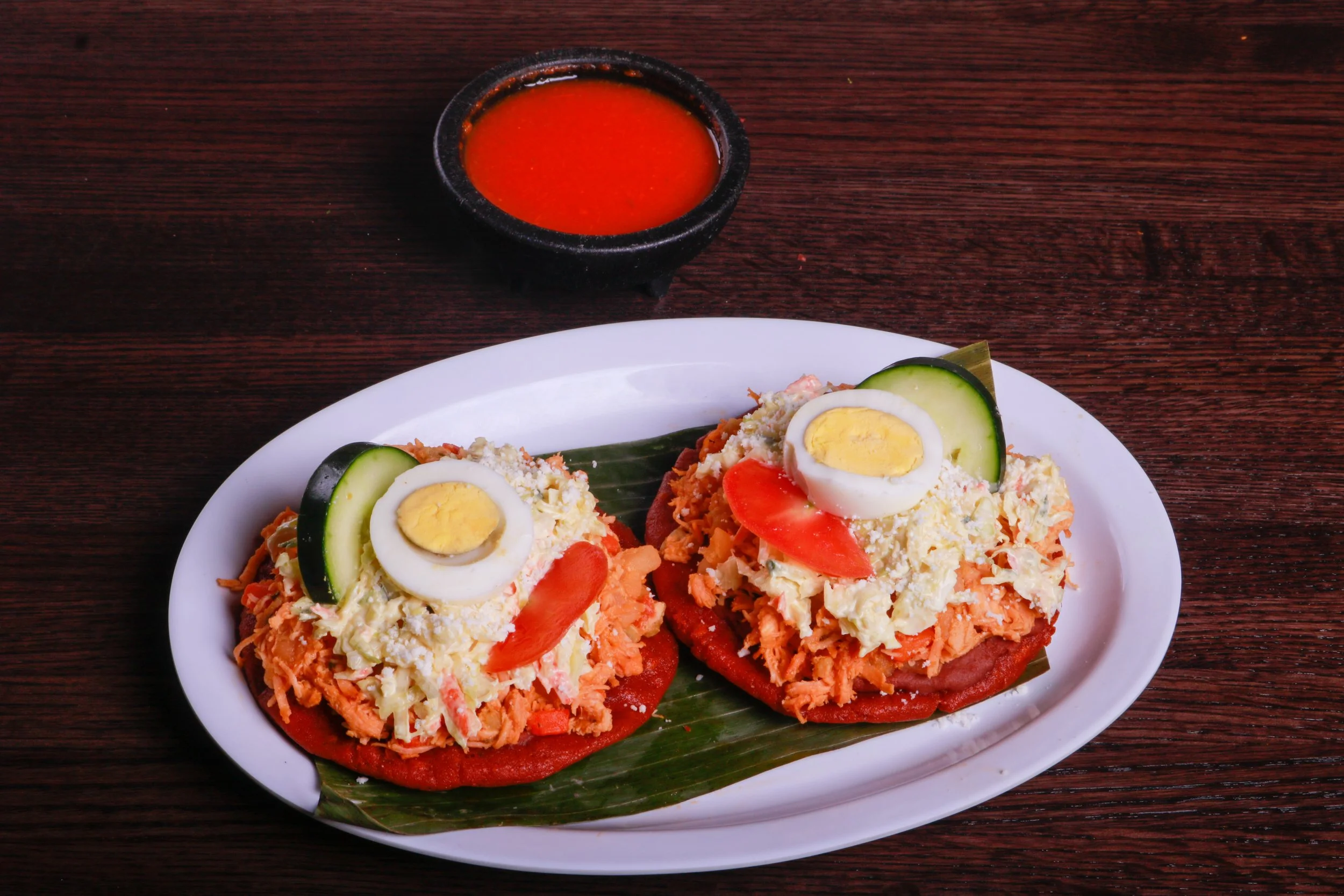 Two colorful tostadas topped with shredded chicken, coleslaw, tomato, cucumber, and sliced hard-boiled eggs, served with red salsa