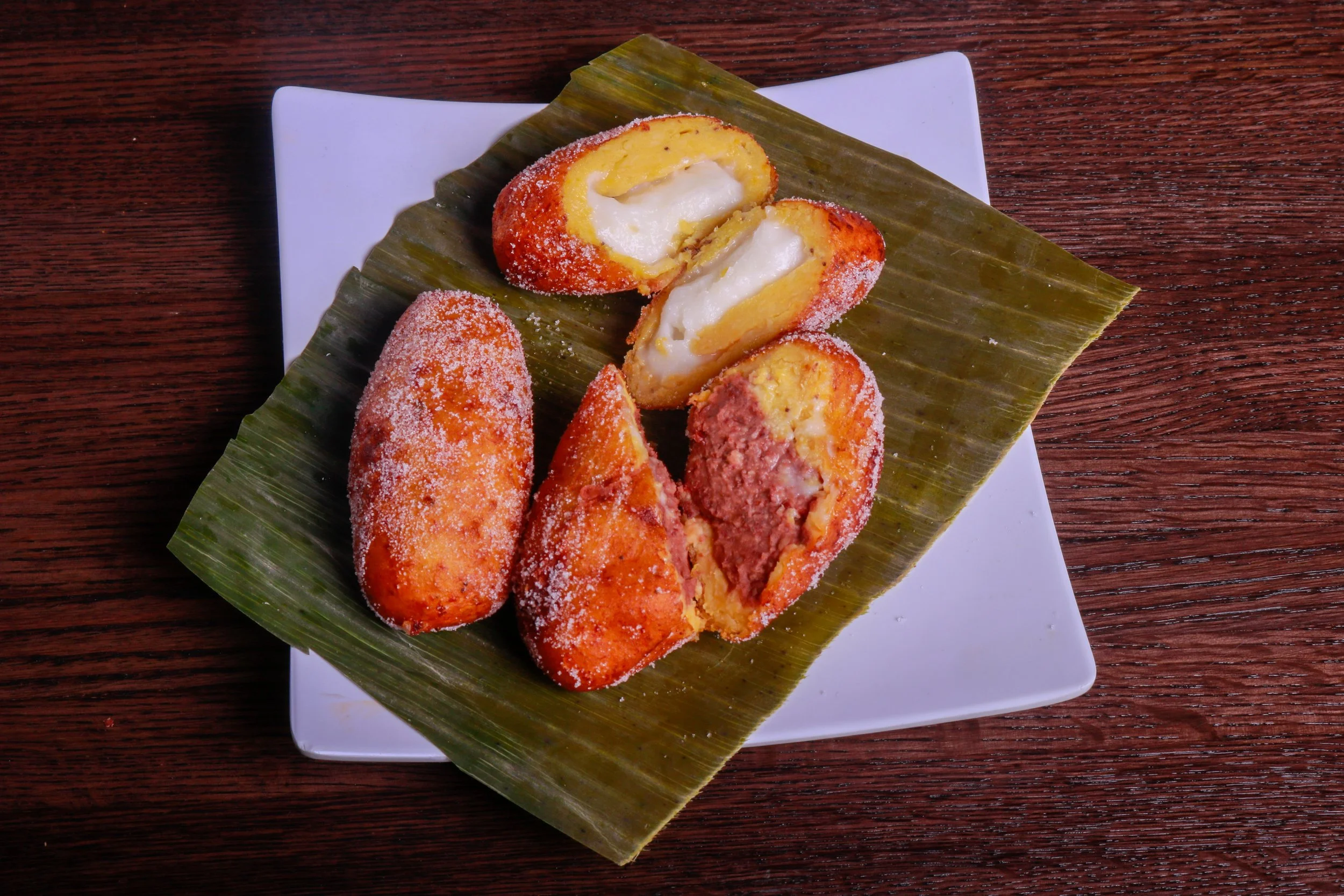 Assorted golden fried pastries dusted with sugar, some split open to reveal creamy white and red fillings, served on a banana leaf-lined plate.