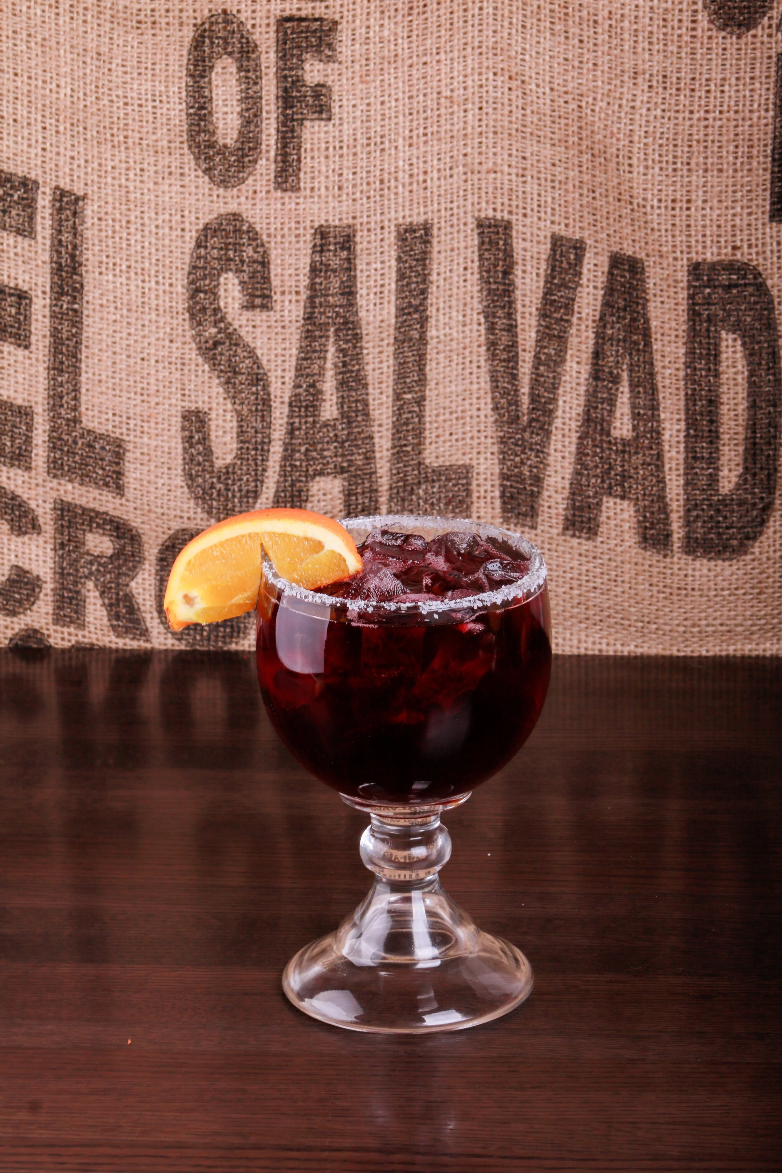 Dark red cocktail with ice and a salted rim, garnished with an orange slice in a stemmed glass