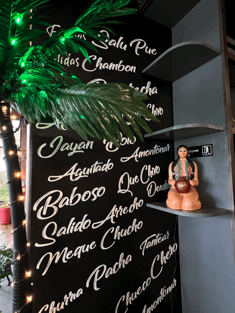 Restaurant display with tropical leaves, Spanish script lettering, string lights, and a small seated woman figurine on shelves