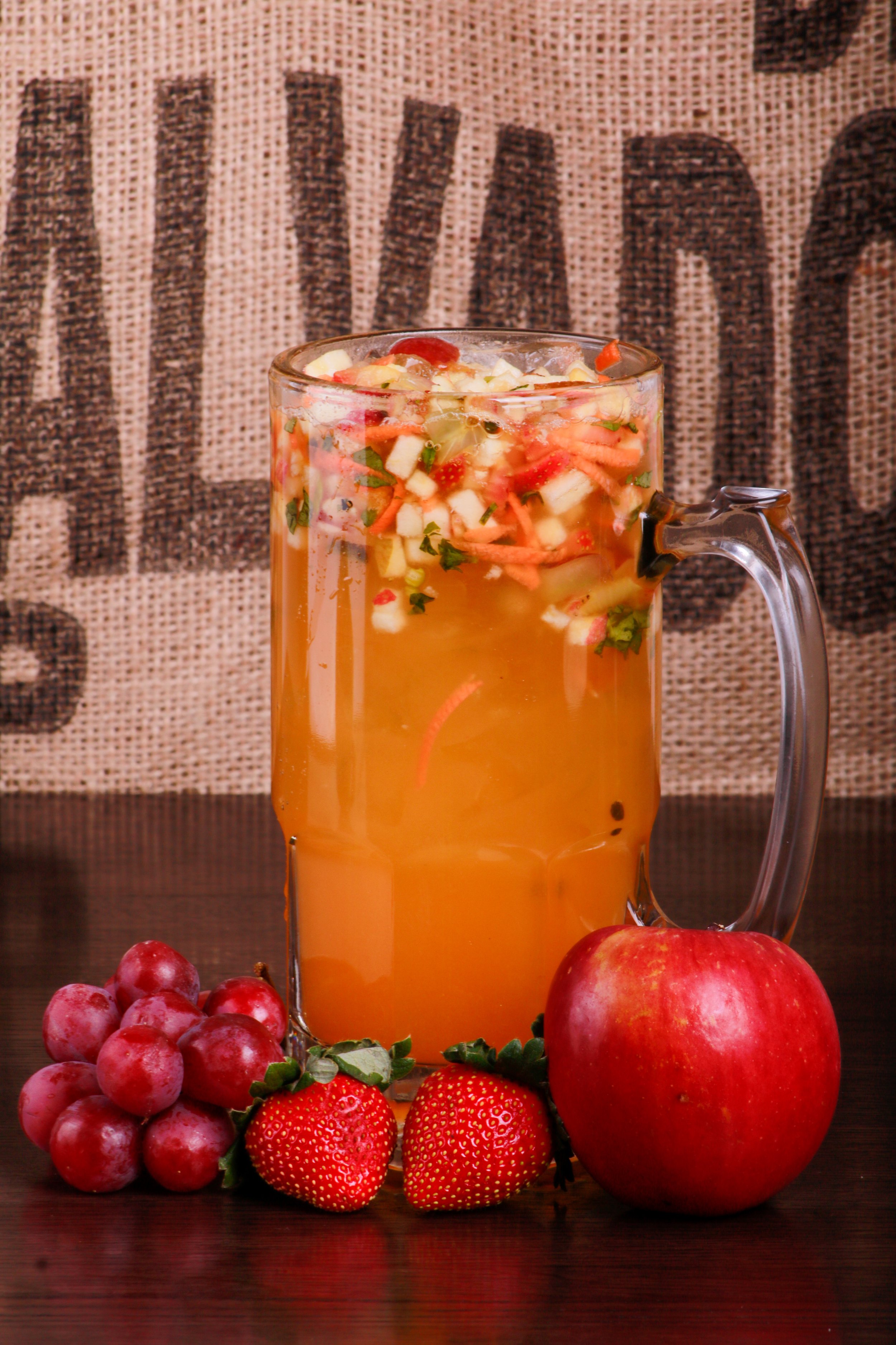 Glass pitcher of orange fruit punch with chopped fruit and mint, surrounded by strawberries, plums, and an apple.