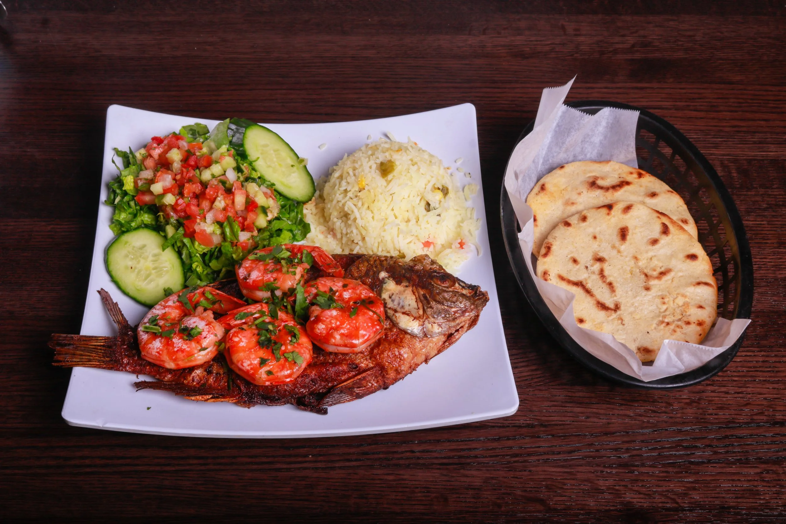 Grilled fish topped with tomatoes and herbs, served with rice, chopped salad, cucumber, and flatbread