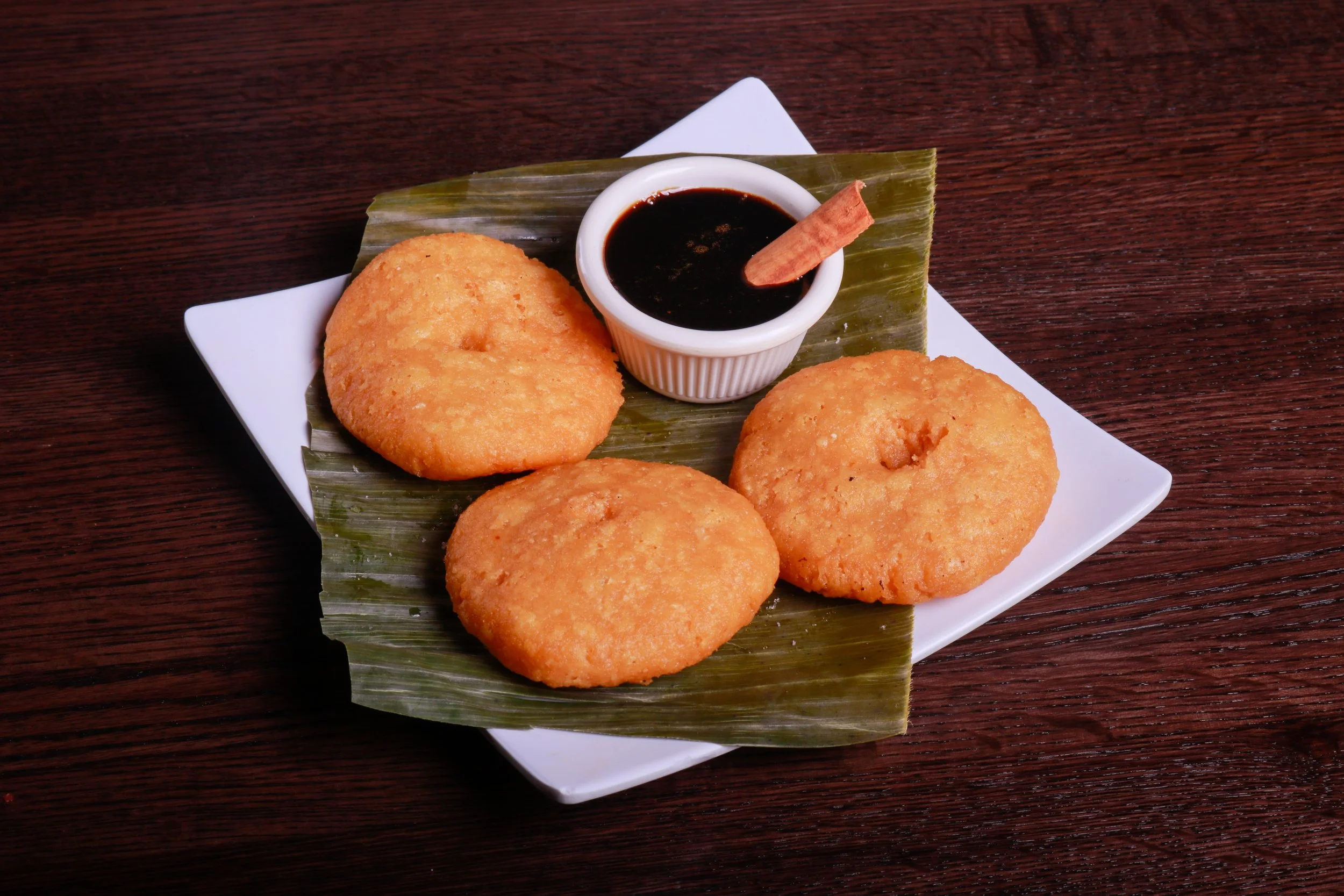 Three golden fried pastries served on a banana leaf with dark dipping sauce and cinnamon sticks