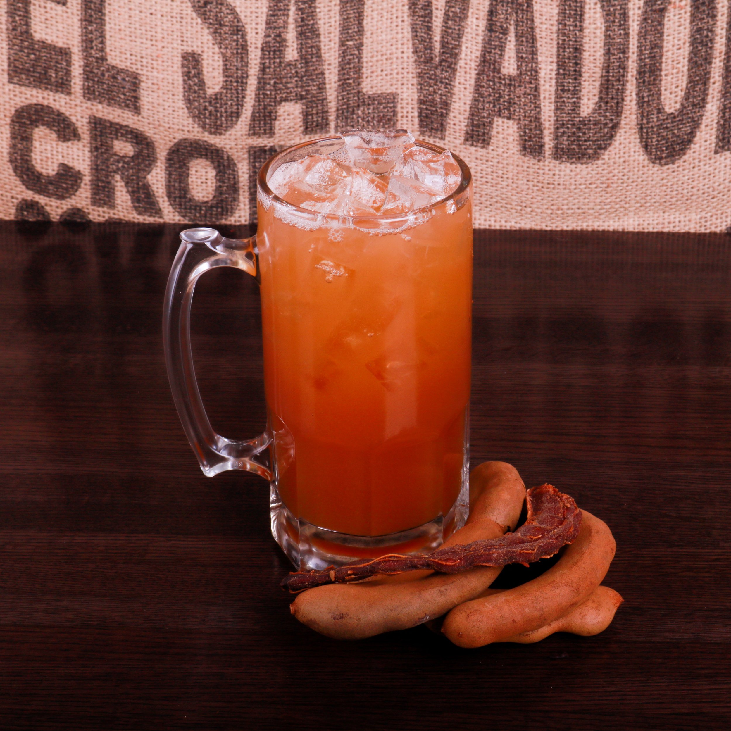 Glass mug of iced orange tamarind drink with tamarind pods on a dark wooden table