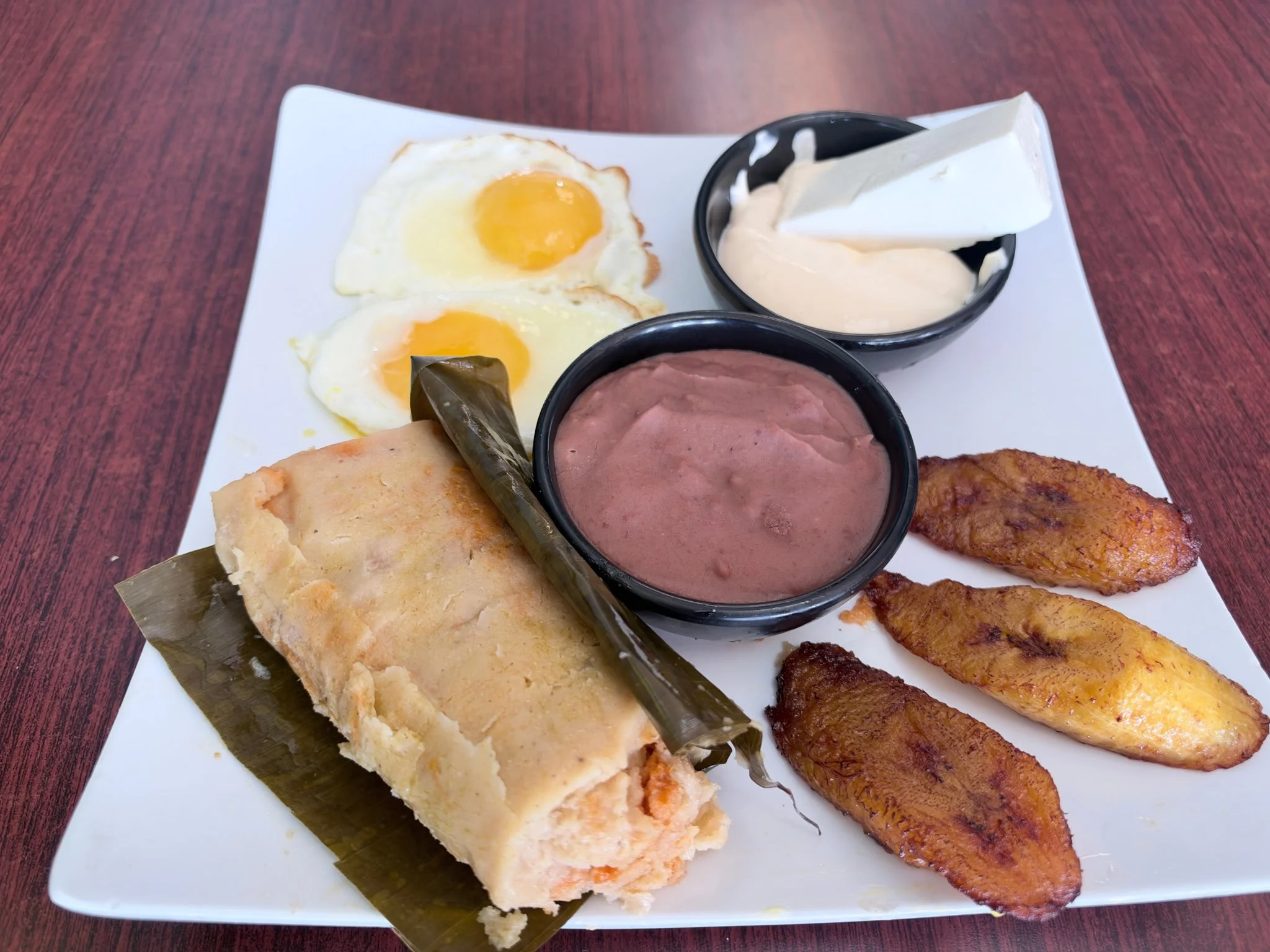 Latin American breakfast platter with fried eggs, plantains, tamales, beans, and cream on a white plate