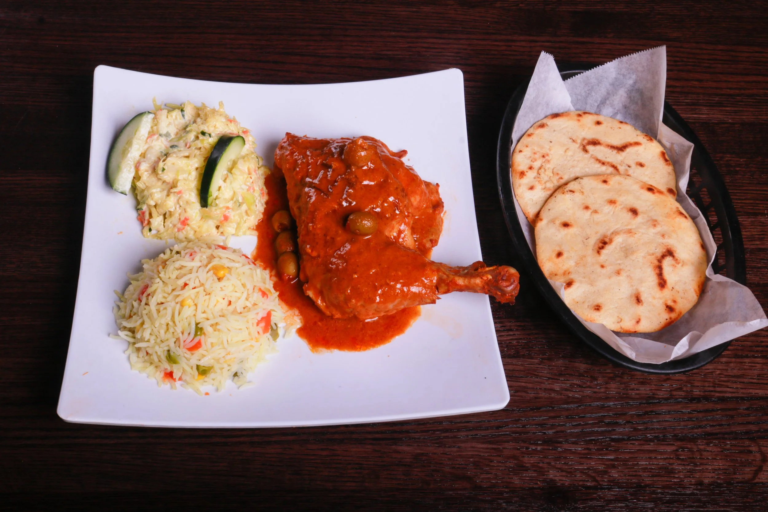 Plate of saucy roasted chicken with olives, vegetable rice, creamy salad, and grilled flatbreads on the side