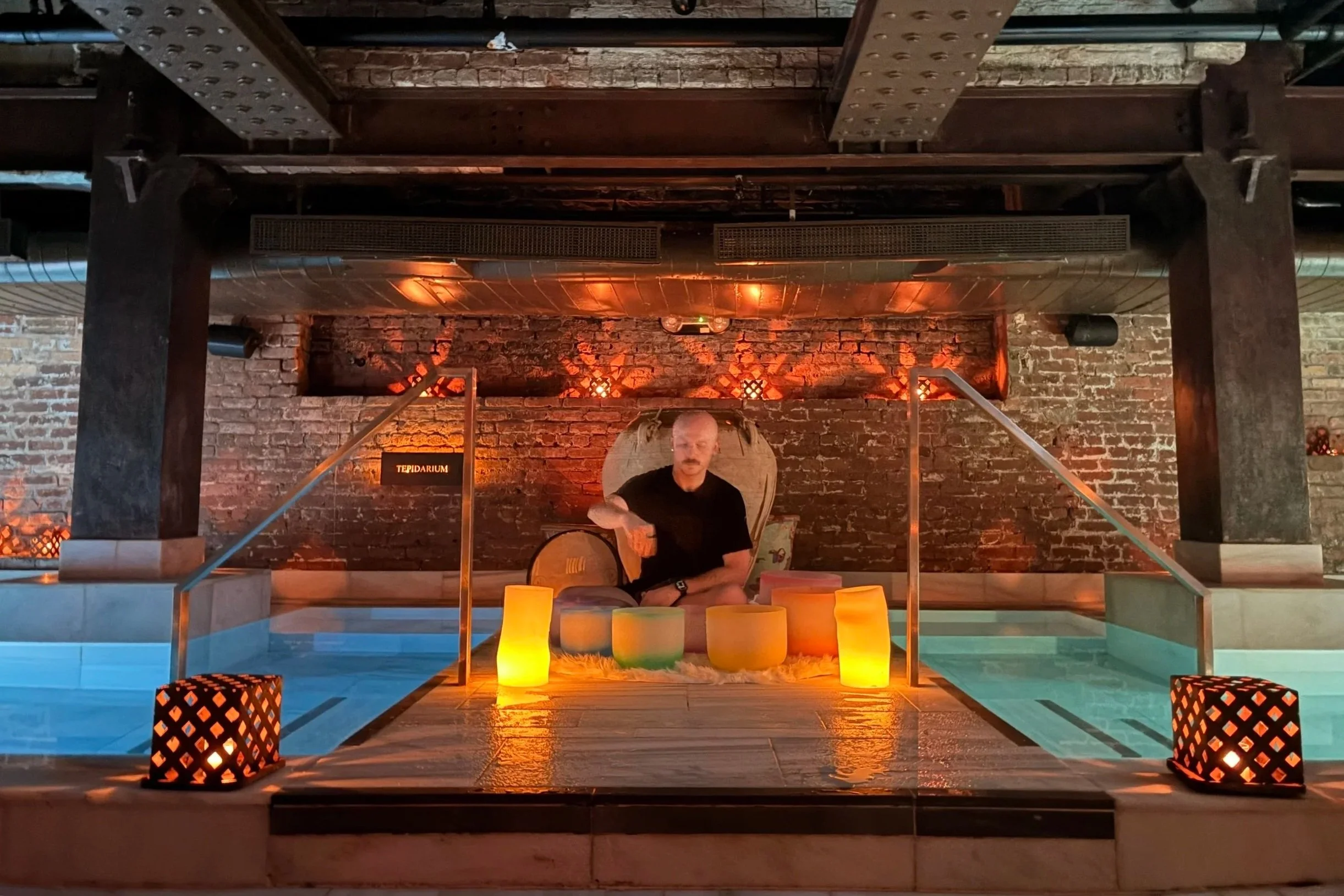 Kyle playing crystal singing bowls in a candlelit indoor spa pool beneath exposed brick and industrial beams