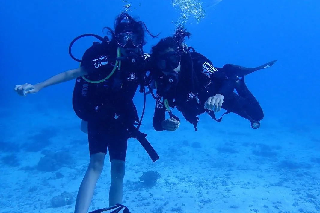 Two scuba divers underwater, holding onto each other, with clear blue water and a sandy ocean floor in the background.