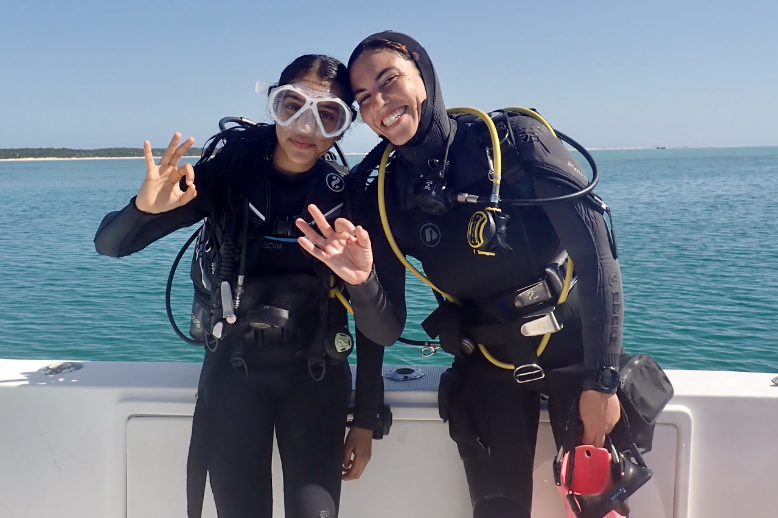 Two people in scuba gear on a boat, smiling with ocean and clear sky in the background.