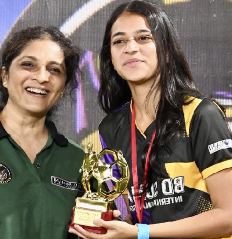 Two women, one older and one younger, smiling at an award ceremony. The younger woman is holding a small golden trophy resembling a soccer ball, wearing a black and yellow sports uniform. The older woman is wearing a green shirt.