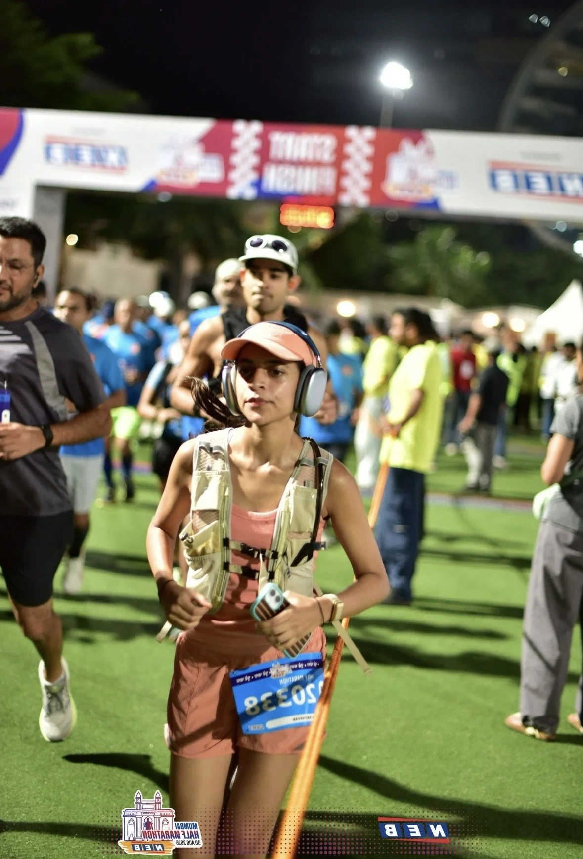 A woman wearing a pink cap, headphones, and a vest, participating in a night race at the Guam Maratón with a race bib number 2038, surrounded by other runners and spectators under bright stadium lights.