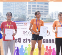 Three women standing on a winners' podium holding certificates and medals, smiling with city buildings in the background.