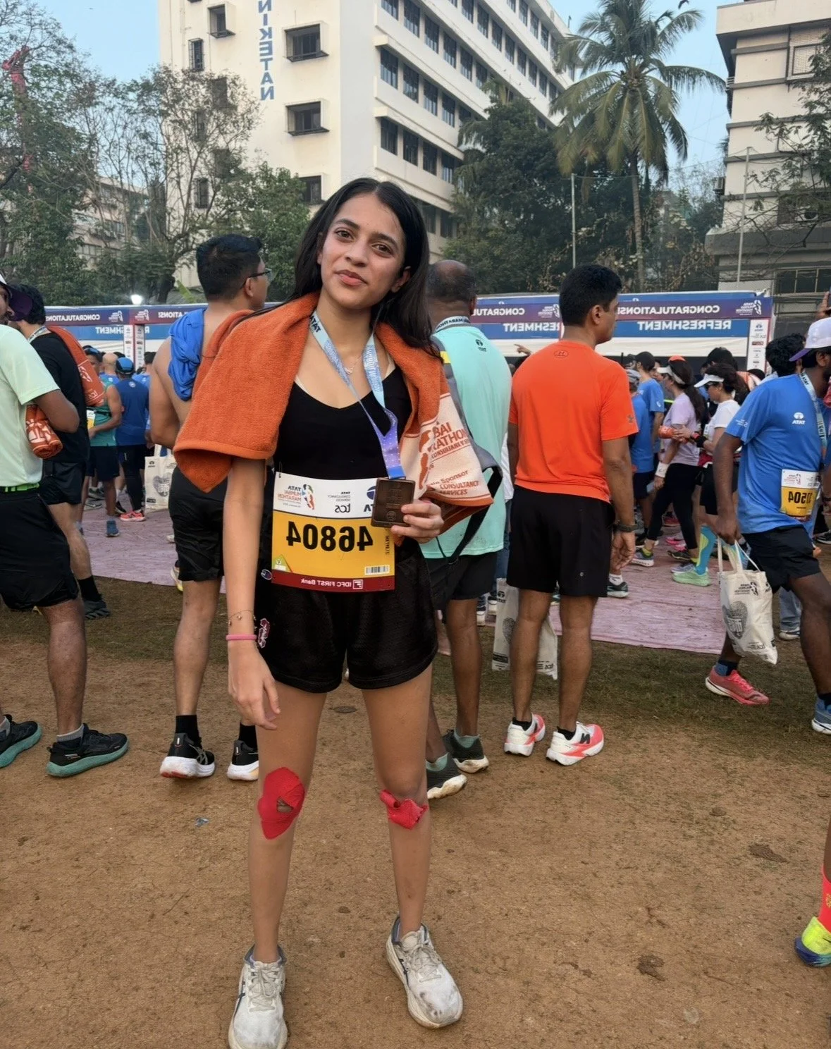 A young woman standing in front of a crowd of runners at a marathon event, wearing running gear, a race bib, medals, and a towel draped over her shoulders. She has knee supports on both legs and is smiling.