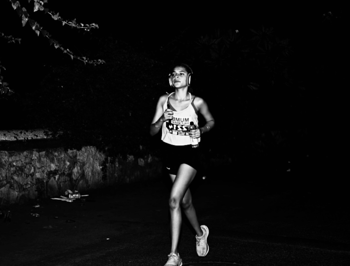 A woman runs at night, wearing headphones, a tank top, and shorts, holding a water bottle.
