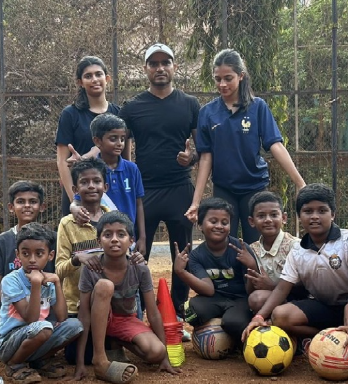 Group of children and an adult posing outdoors on a sports field, with soccer balls, cones, and children in sports uniforms.