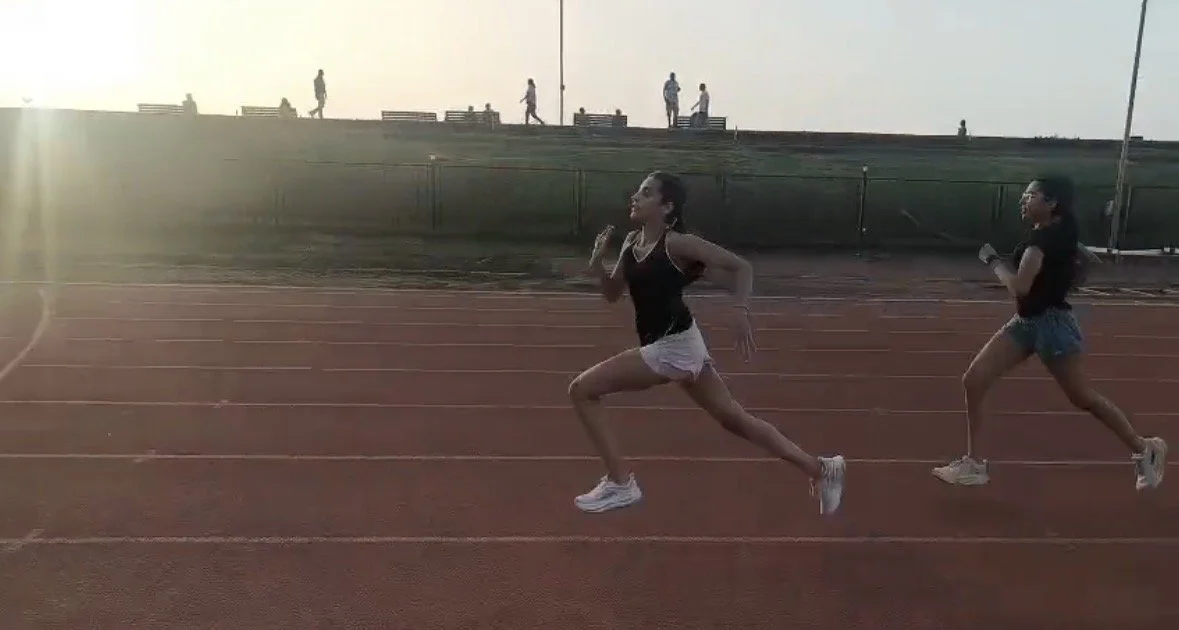 Two women are running on a track during sunset, with several people walking on the grass hill in the background.