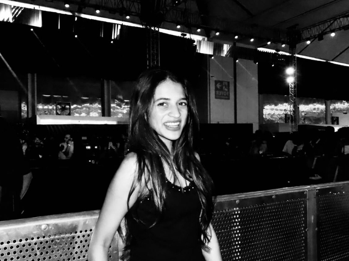 A young woman with long dark hair, smiling, standing in an indoor venue with a stage and audience in the background.