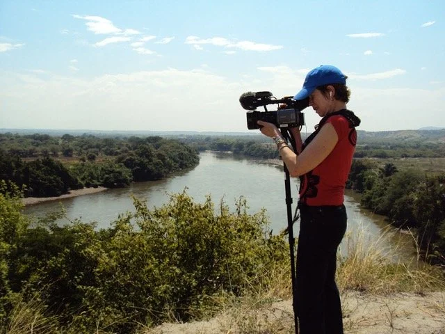 Woman filming with a video camera on a hill overlooking a river and trees.