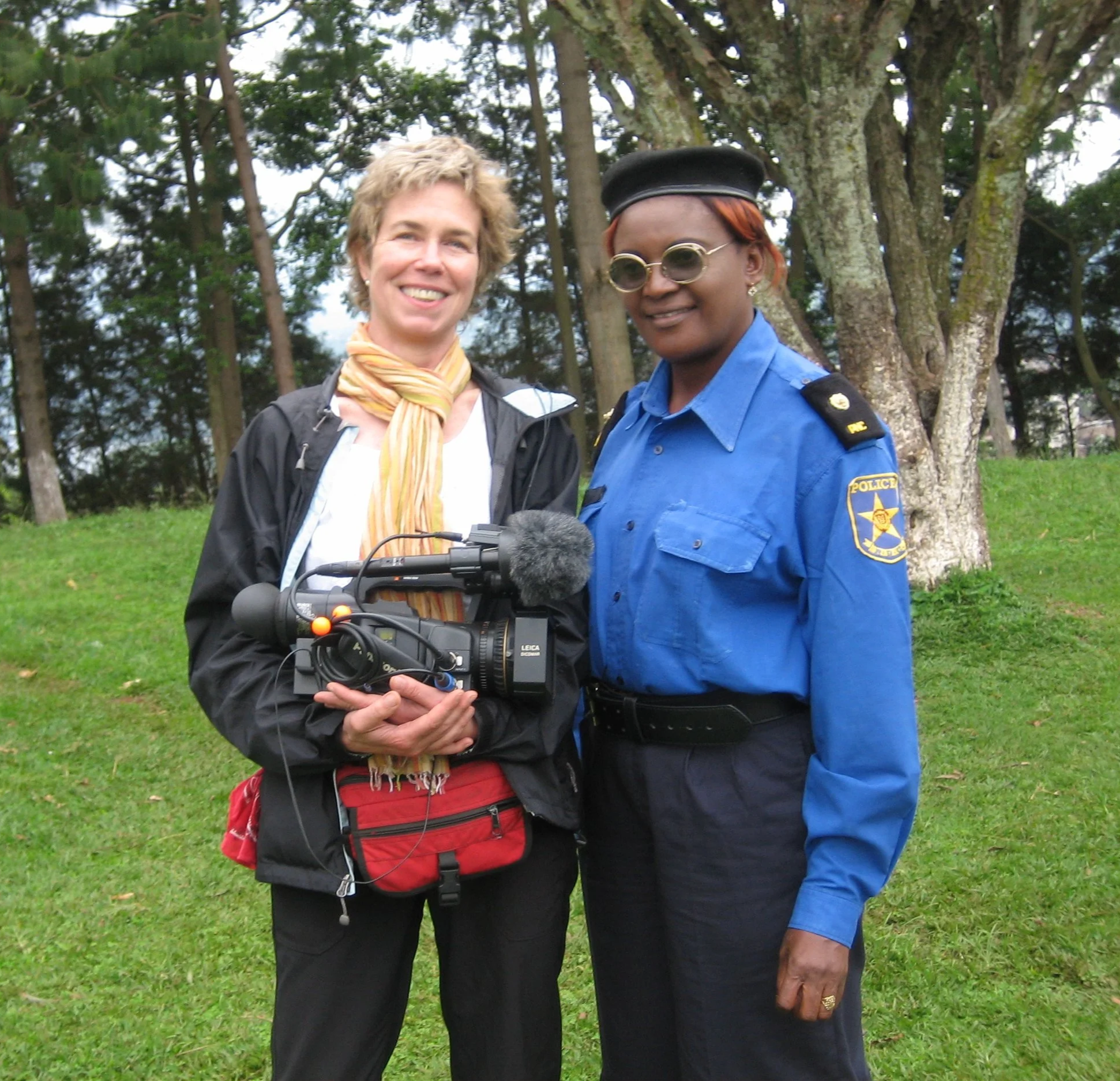 Two women standing together outdoors in a natural setting with trees and grass in the background. One woman is dressed casually with a scarf and jacket, holding a camera, while the other woman is in a police uniform with round sunglasses and a beret.