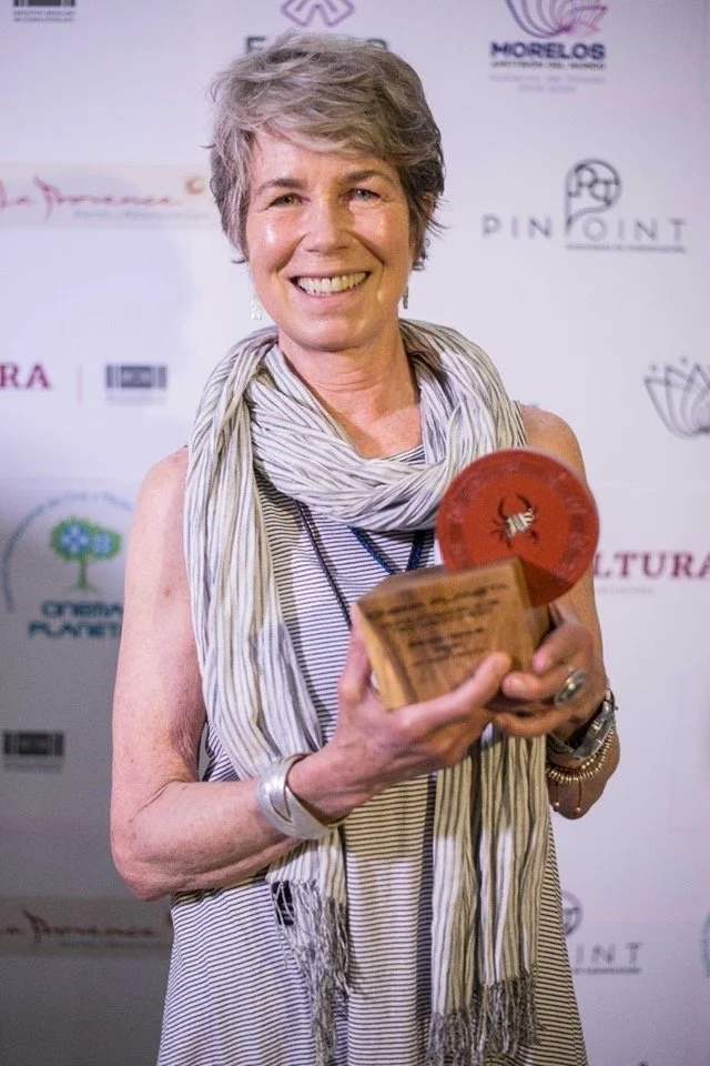 A smiling woman with short gray hair holding a wooden award. She is wearing a striped sleeveless top and a light scarf, standing in front of a backdrop with various logos.