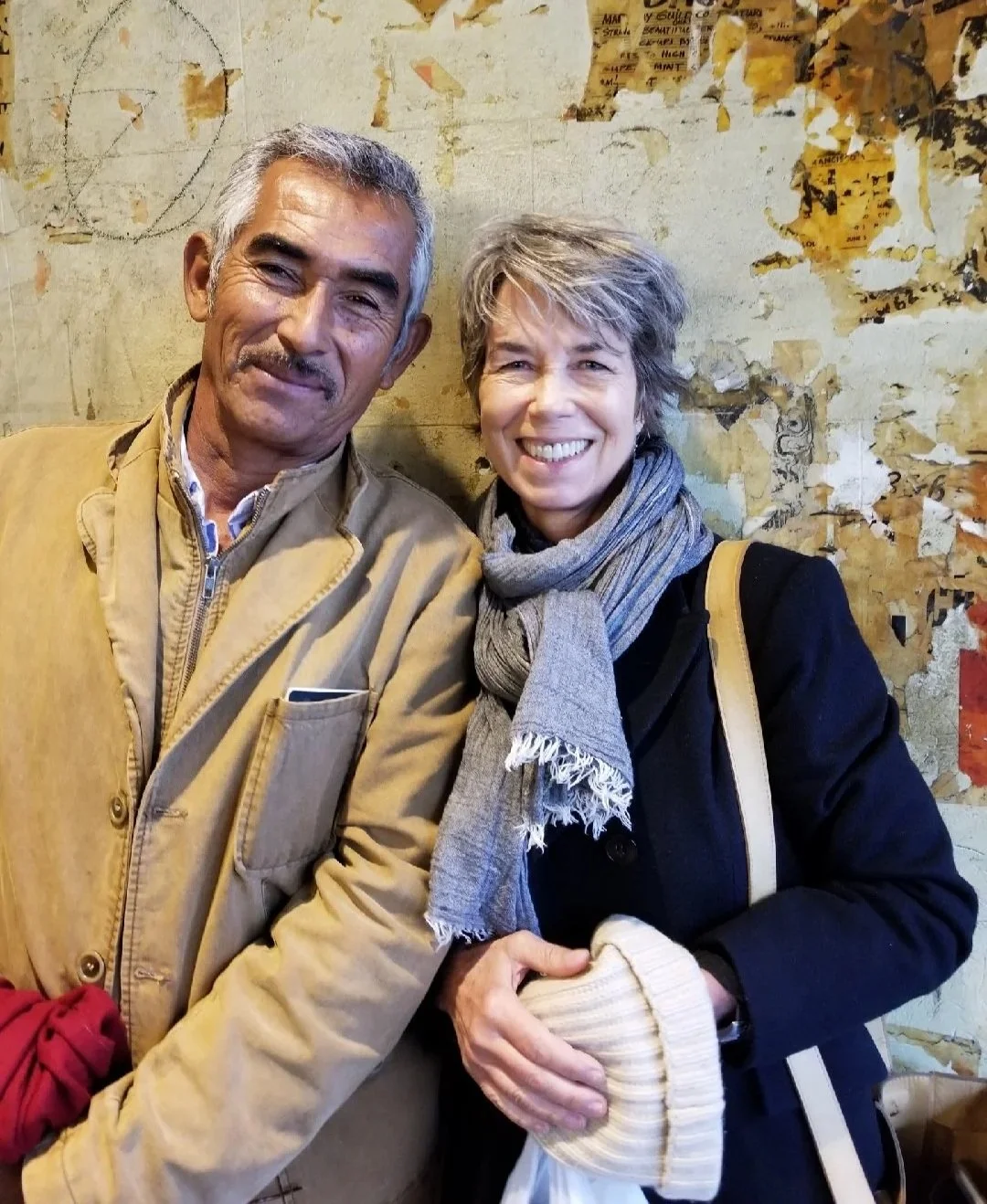 Two smiling people, a man and a woman, standing close together in front of a weathered wall with peeling paint and posters.