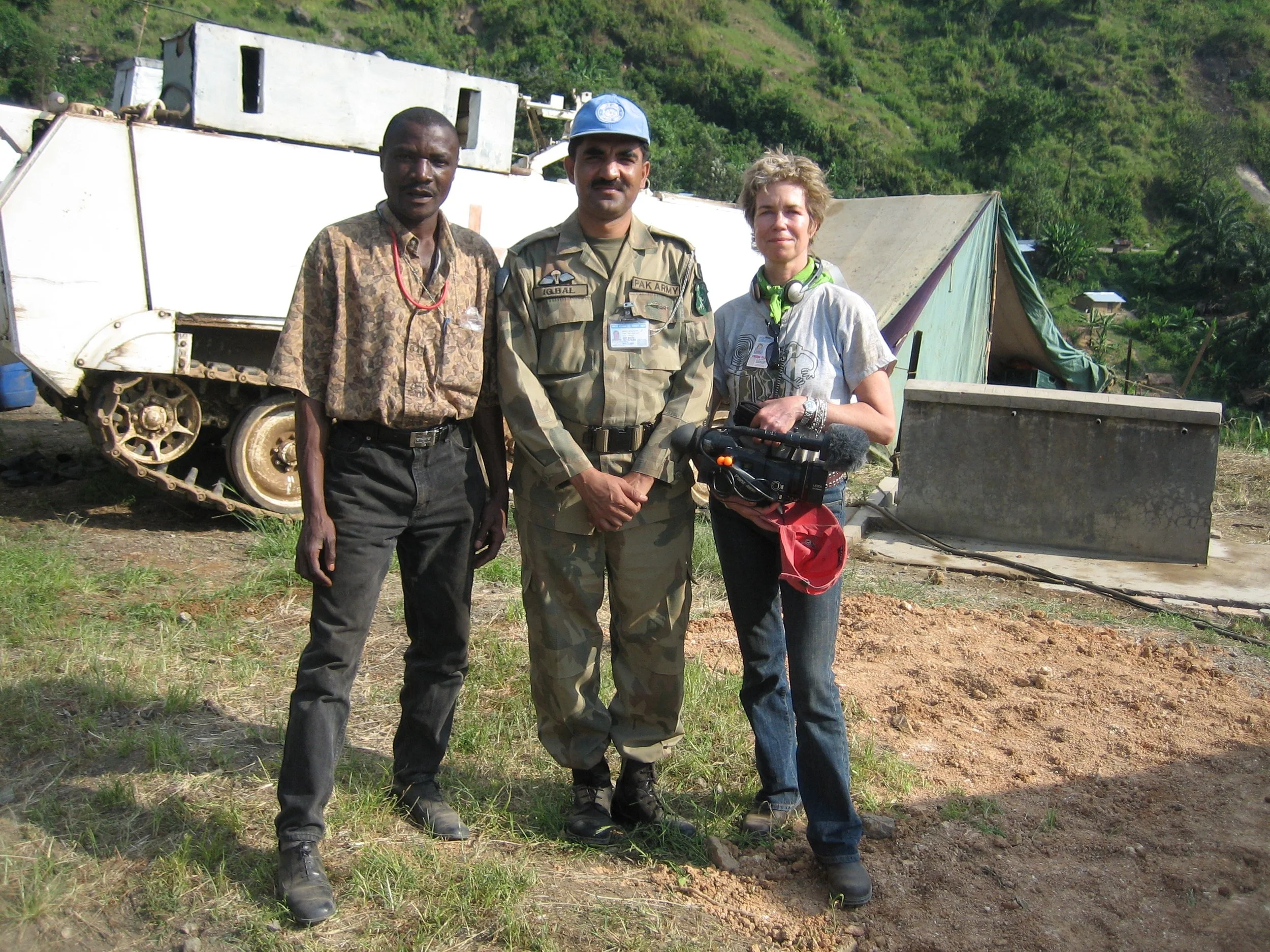 Three people standing outdoors in front of a military vehicle, with green hills in the background. The individuals include a man in military uniform, a woman holding a camera, and another man in casual clothing.