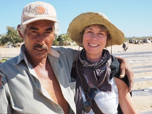A man and a woman smiling and posing together outdoors, with the man wearing a baseball cap and the woman wearing a wide-brimmed hat and bandana, in a sunny environment with horses in the background.