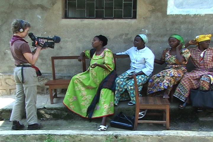 A woman, Lisa Jackson,  with headphones filming four women sitting on a bench outside.