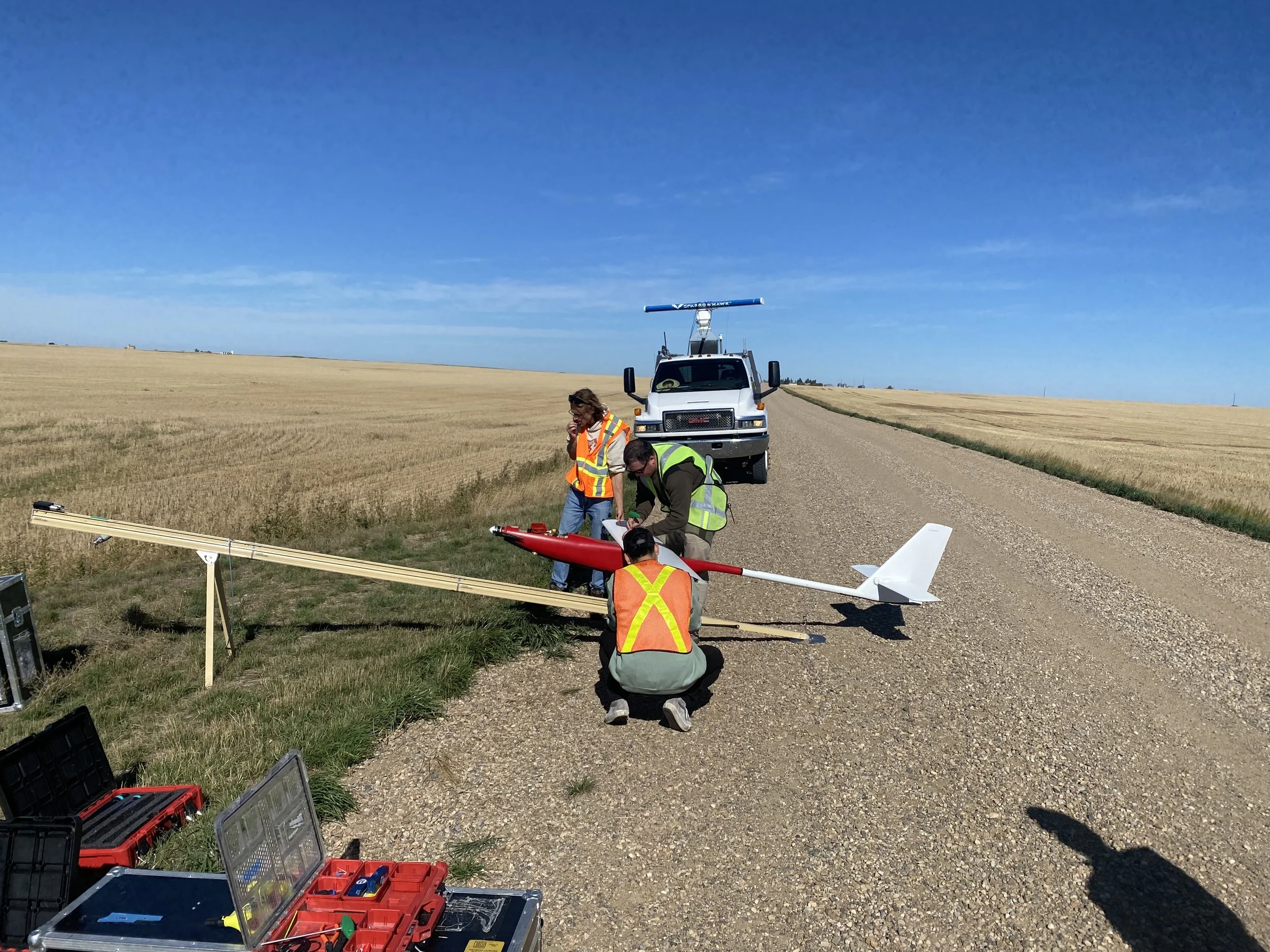 Survey crew assembling a small unmanned aircraft beside a truck on a rural gravel road surrounded by harvested fields.