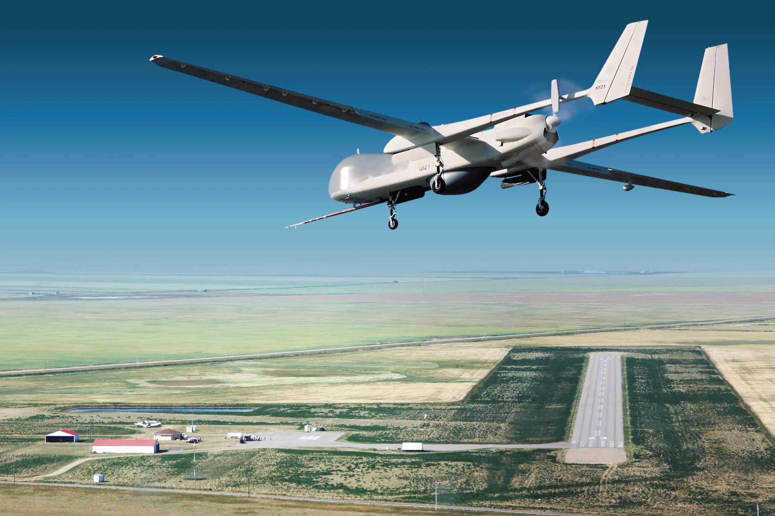 White military drone flying over a rural airfield and runway under a clear blue sky