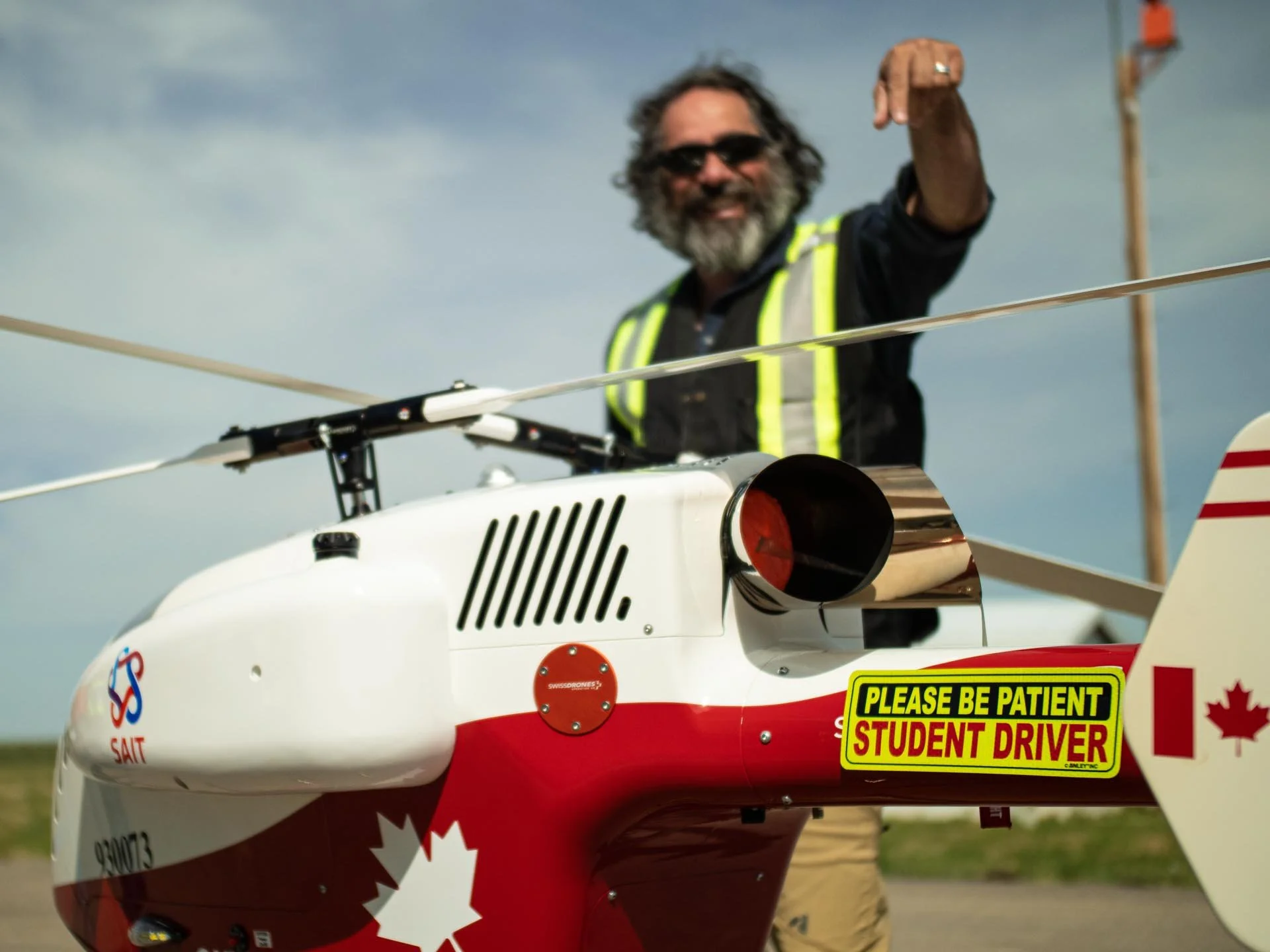 Flight instructor in a high-visibility vest standing behind a red-and-white student training aircraft with a 'Please Be Patient Student Driver' sign and Canadian flag decals