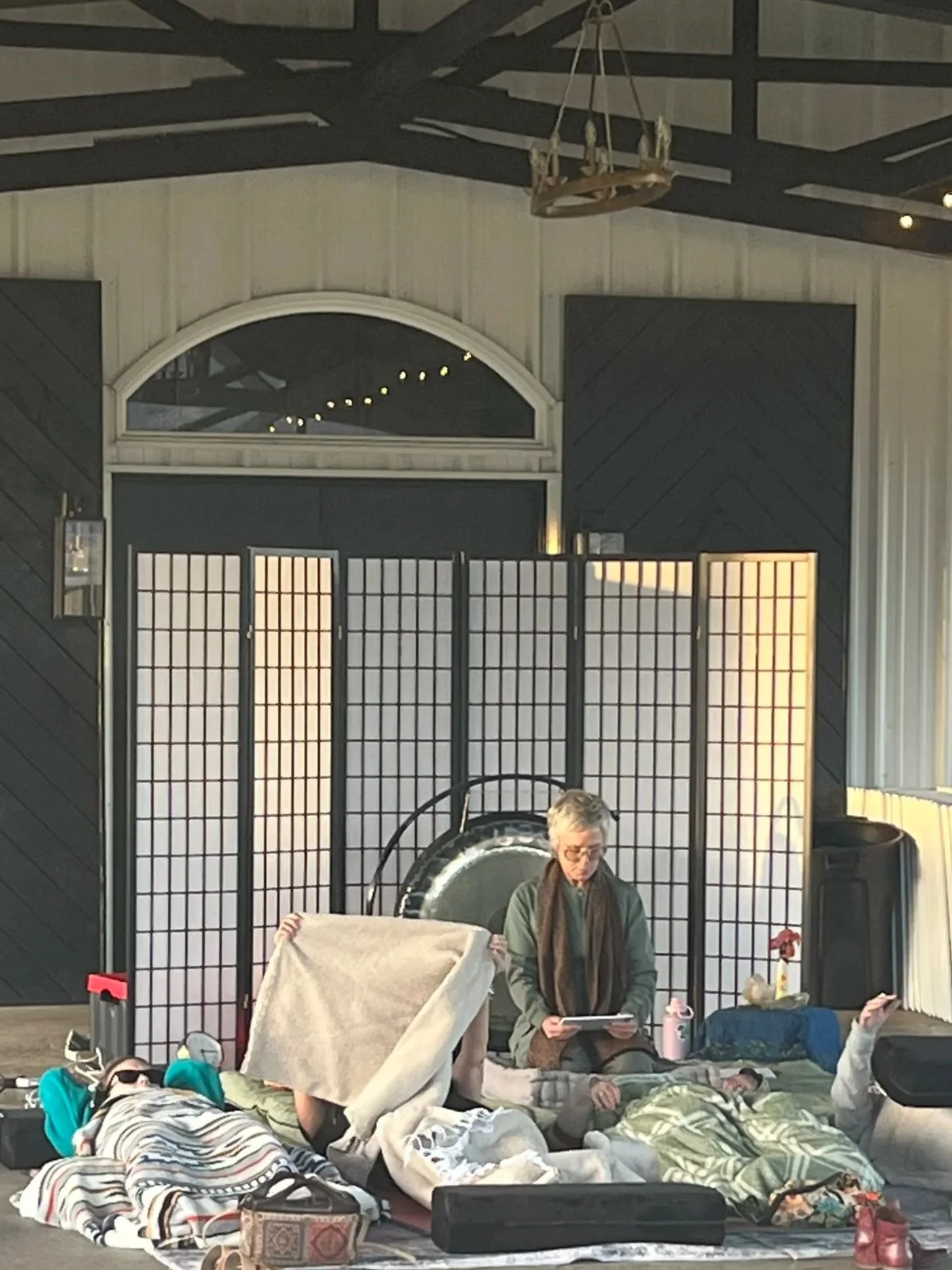Group of people lying on the floor with blankets, and a woman sitting up reading a book in front of a black folding screen, with a gong and other objects in the background.