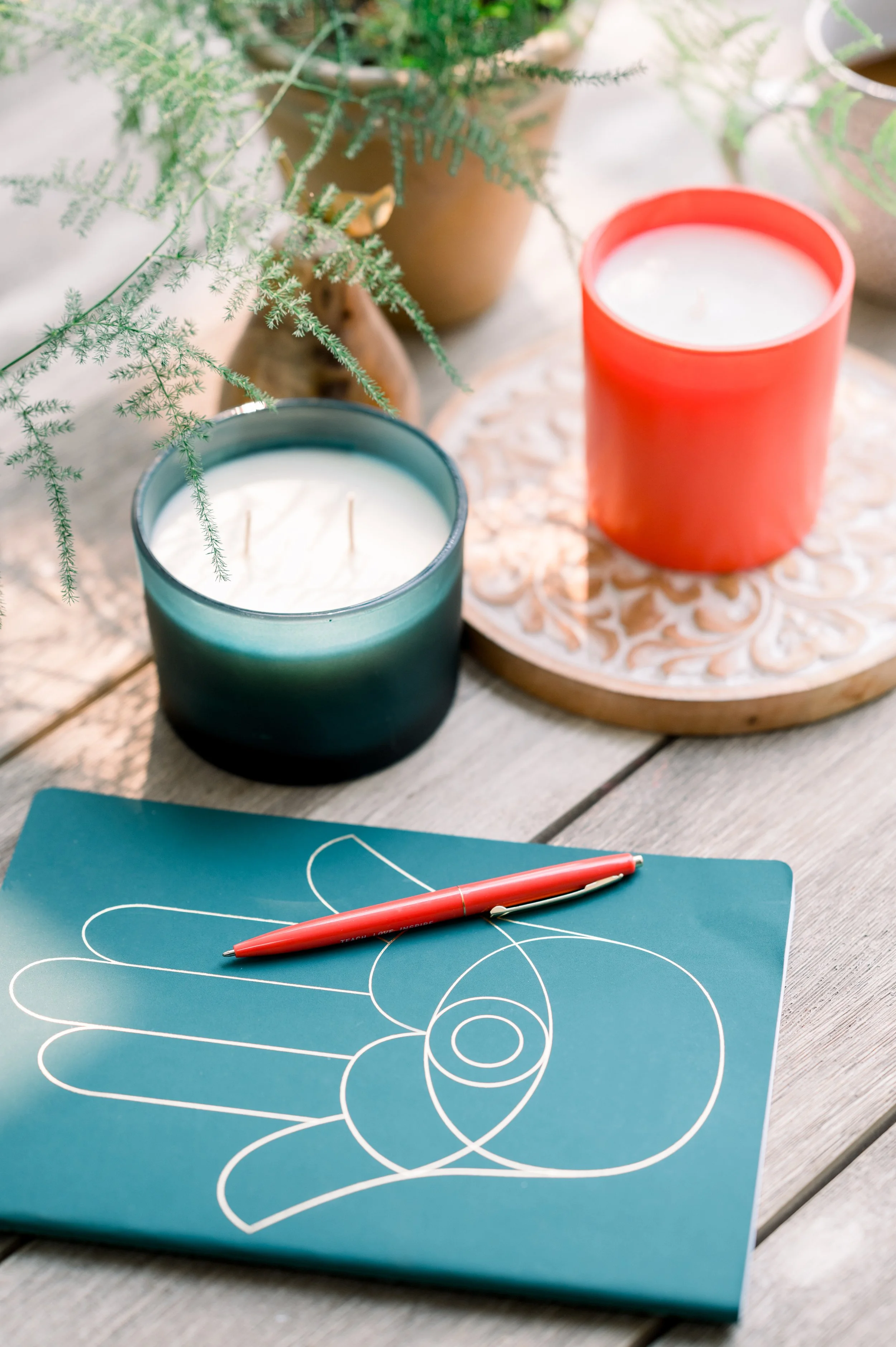 Close-up of a blue notebook with white spirals drawn on it, a red pen resting on top, and candles in teal and red holders. Potted greenery is blurred in the background.
