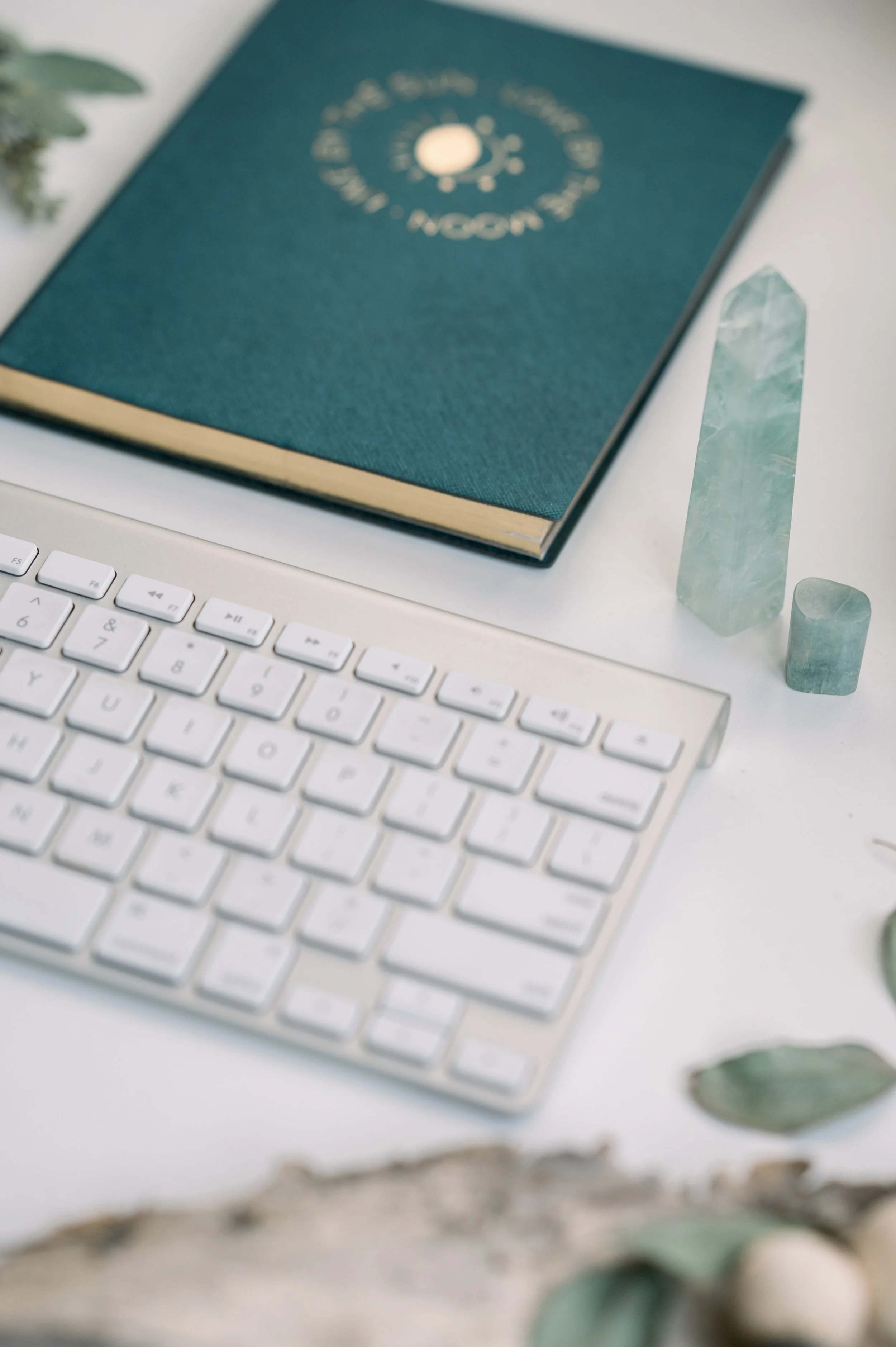 A white computer keyboard, a teal hardcover book, and decorative green crystal stones on a white surface.