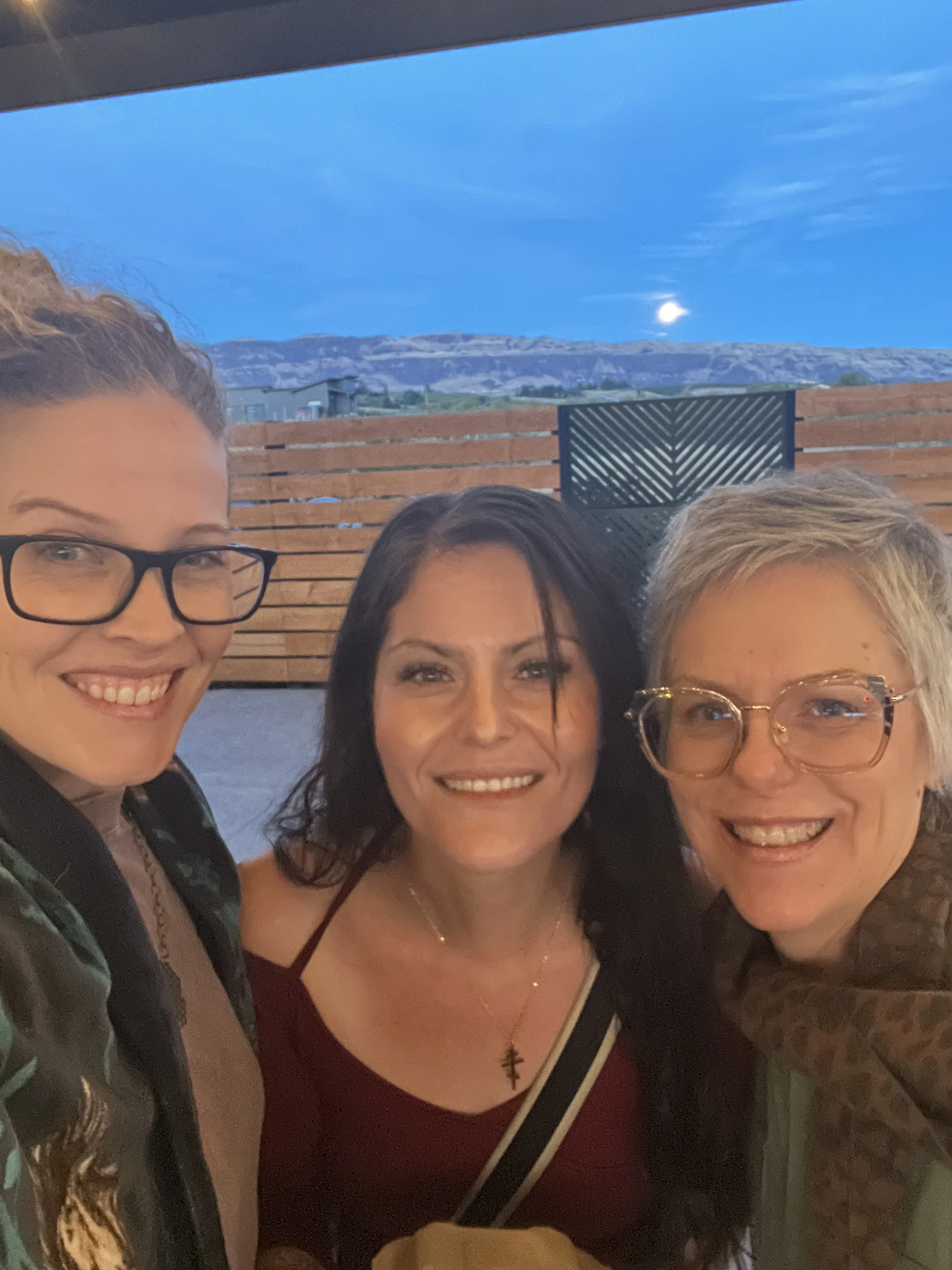 Three women taking a selfie outdoors during the evening, with mountains and a bright moon in the background.