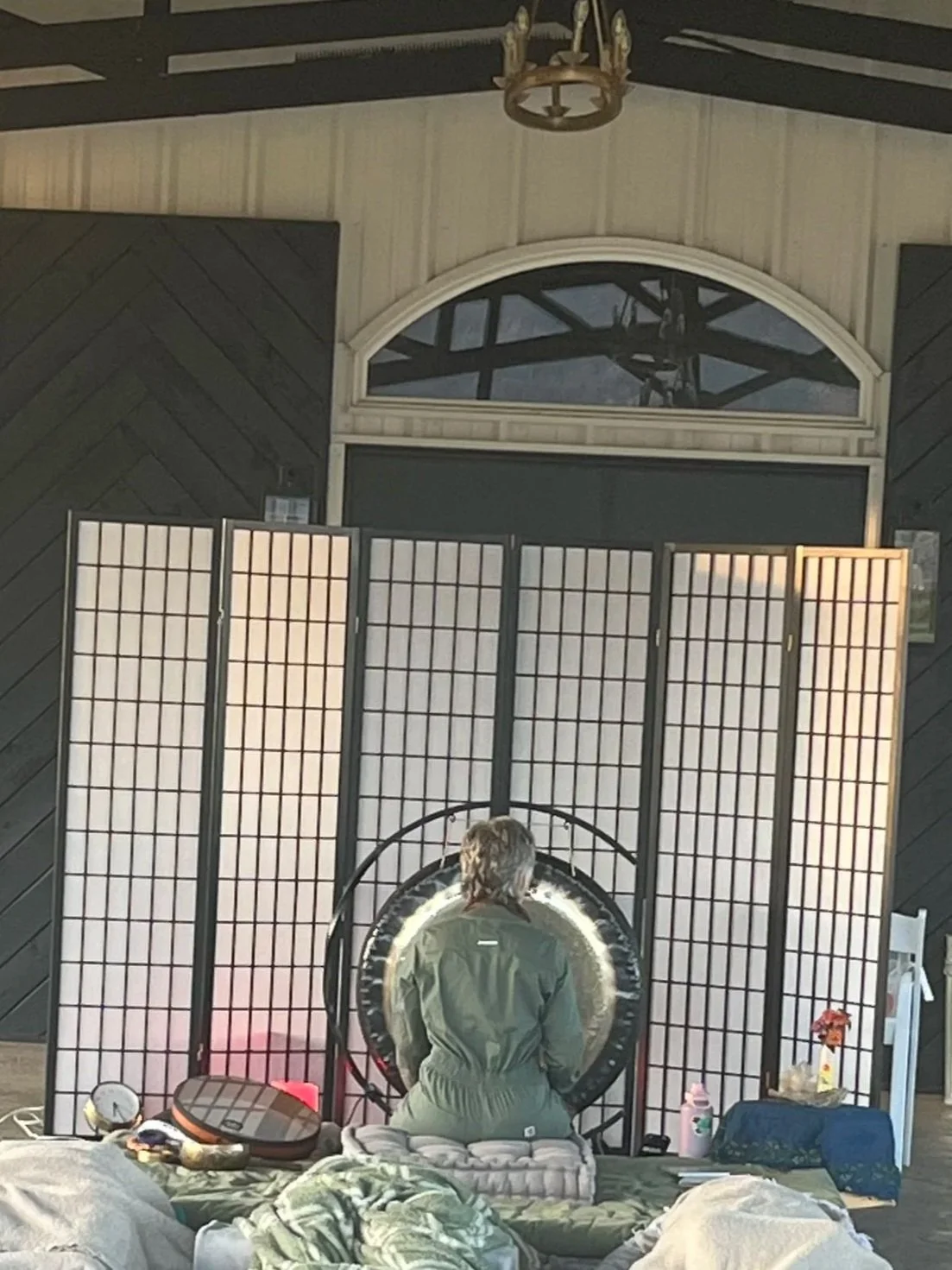 A person with short gray hair sitting on a cushioned bench, facing a large circular gong, in front of a folding screen divided into four sections. The room has dark wooden walls, a window above with a reflective surface, and a chandelier hanging from the ceiling.