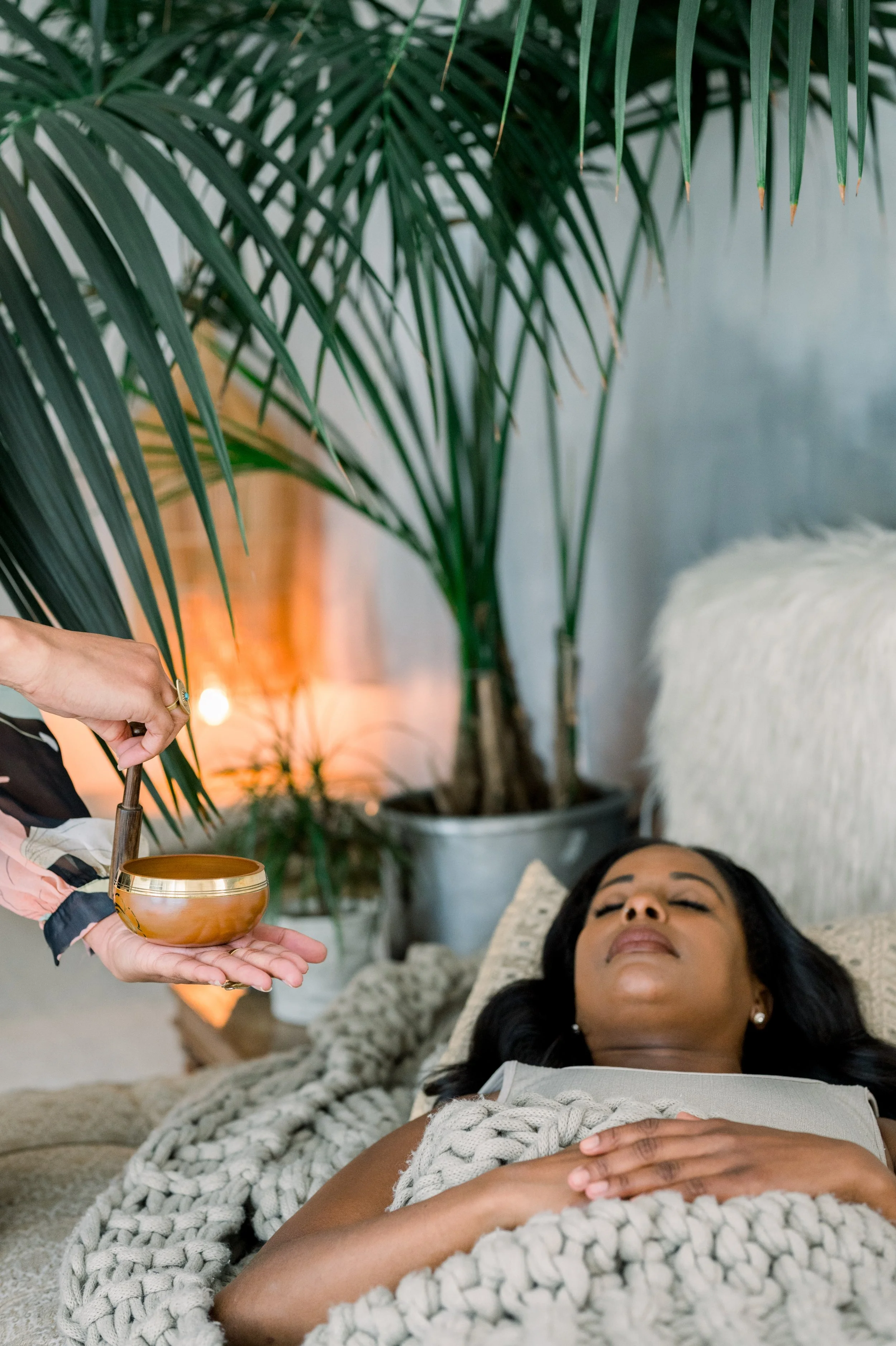 A woman lying on a bed with her eyes closed, covered with a chunky knit blanket, with a large potted plant and a white furry pillow in the background. Someone's hand is holding a wooden bowl and a spoon near her face.