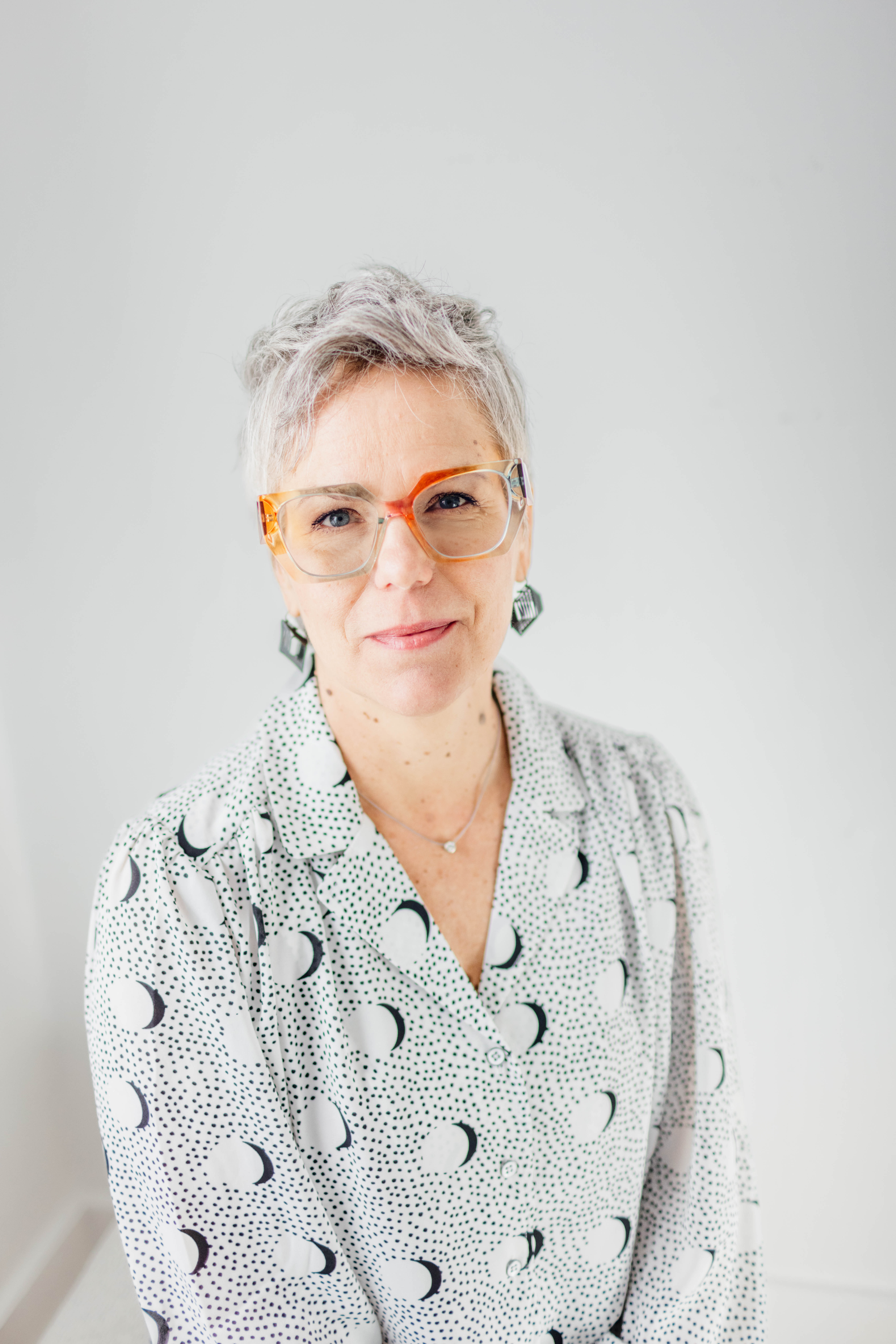 Portrait of a smiling woman with gray hair, translucent orange glasses, geometric earrings, and a patterned blouse