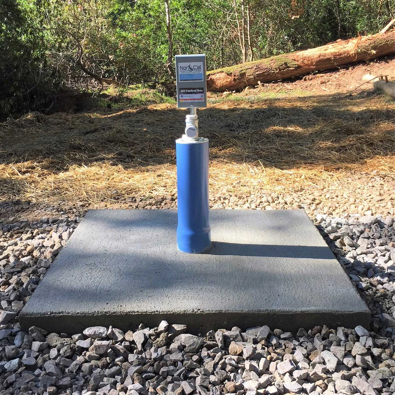 A water control box on a blue pipe stands on a concrete platform outdoors, with a sign on the box labeled 'NorthCal' and 'QDC Control Box.' The surrounding area has gravel and dirt, with trees and fallen logs in the background.