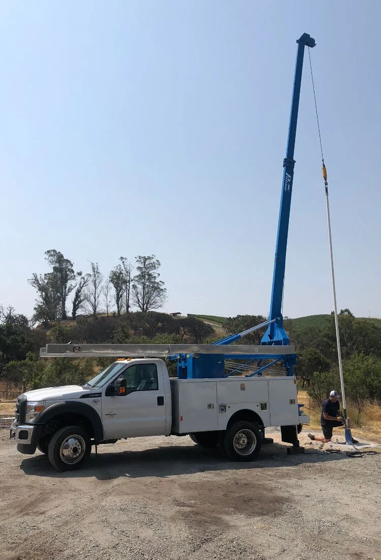 A utility truck with an extended boom lift and a worker adjusting equipment on the ground in an outdoor setting with trees and hills in the background.