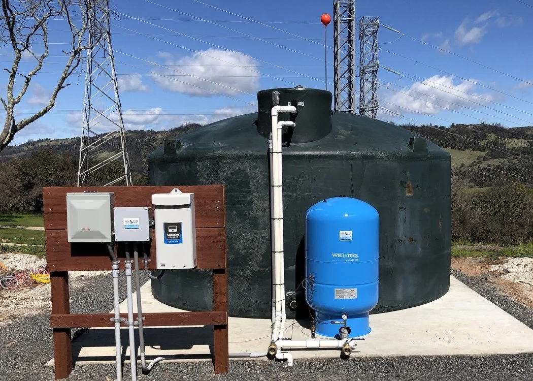 A large black rainwater collection tank with blue pressure tank and filtration system outdoors, set on a concrete pad with a rural landscape in the background.
