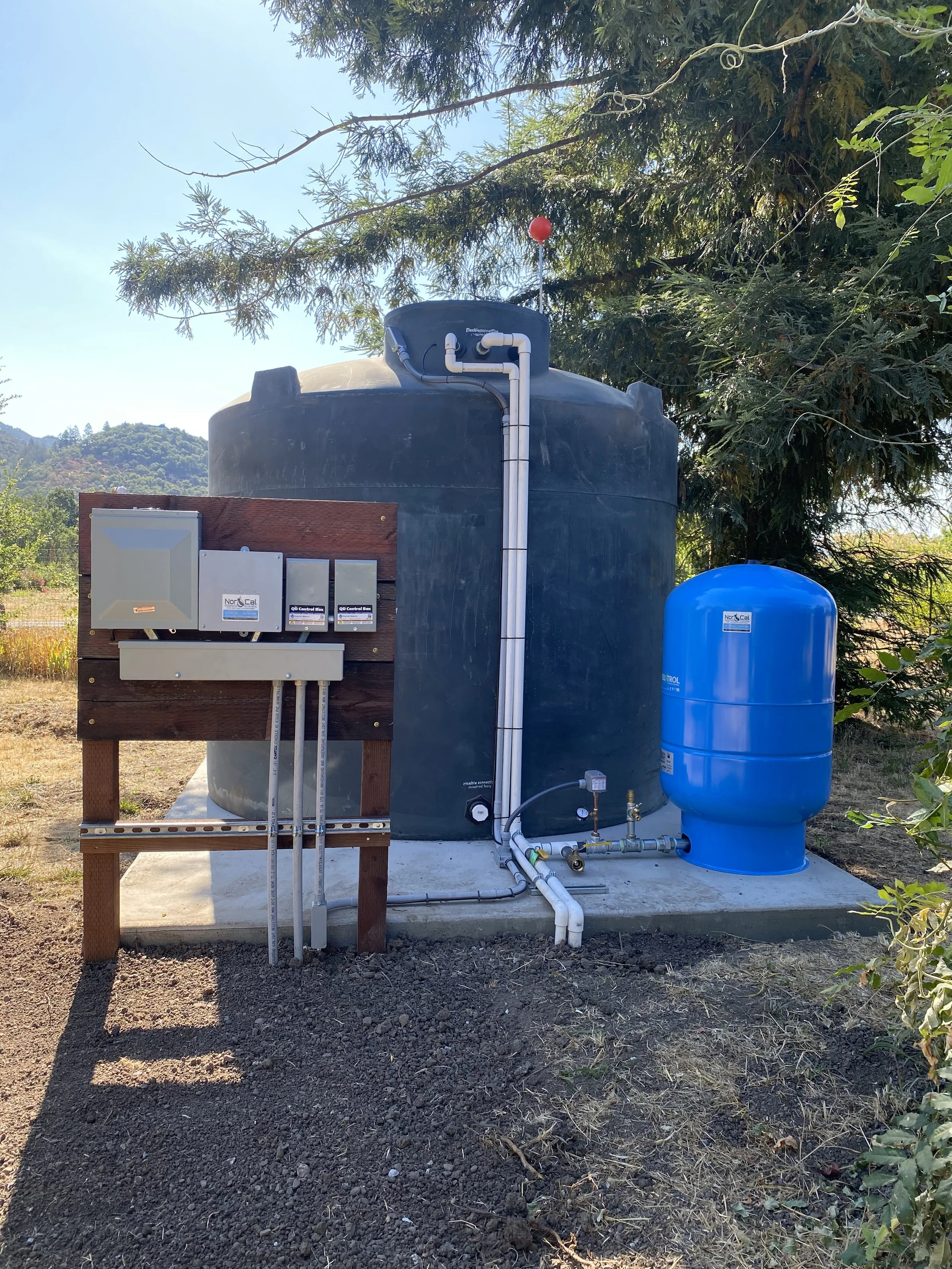 A water treatment or filtration system installed outdoors on a concrete slab, with large blue and black tanks connected by pipes, mounted on a wooden board, surrounded by natural greenery and trees.