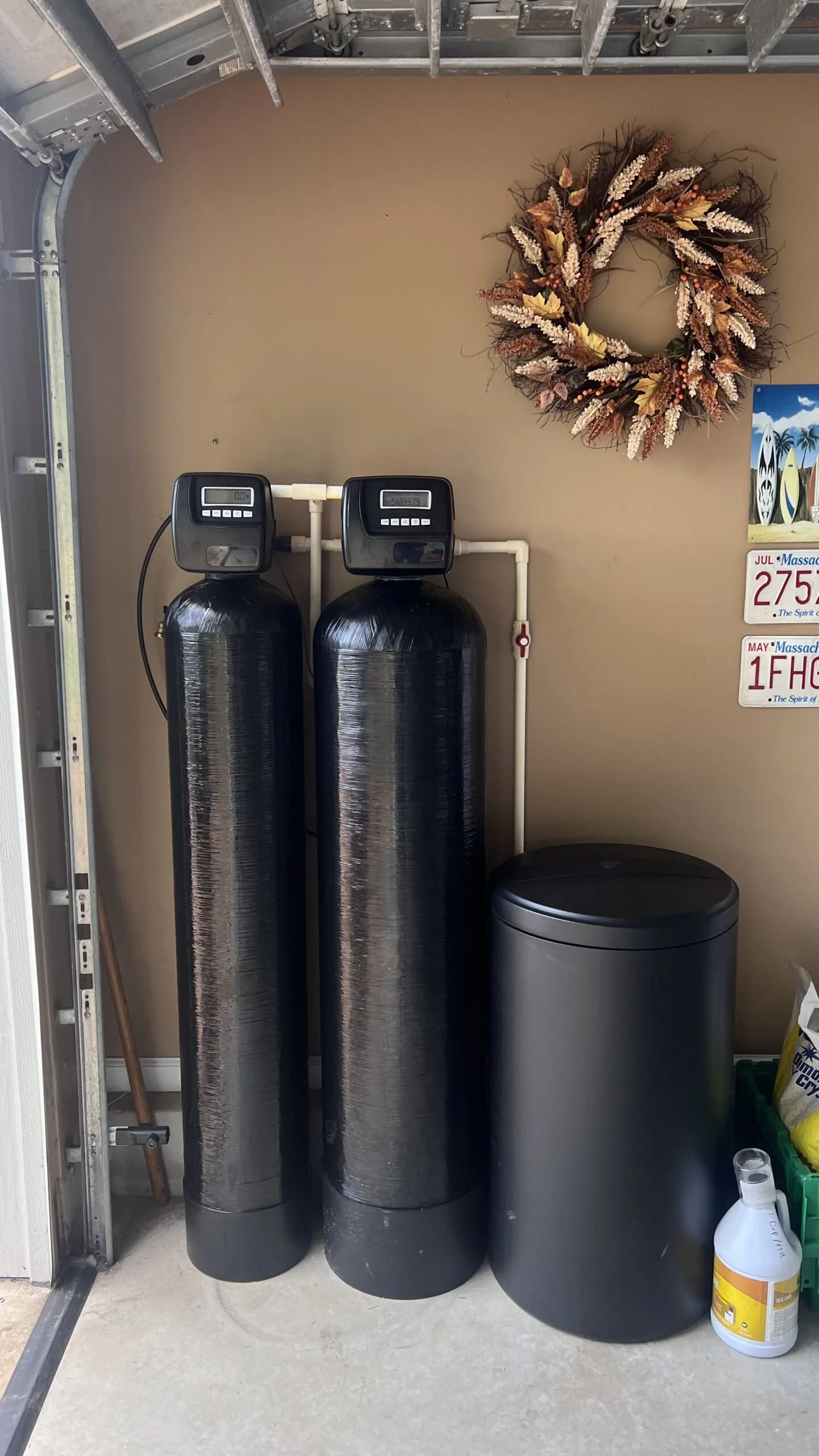 Two large black water softener tanks with digital control heads, a smaller black trash can, and a yellow cleaning product bottle on a concrete floor in a utility area. A decorative autumn wreath hangs on the beige wall in the background, along with some license plates.