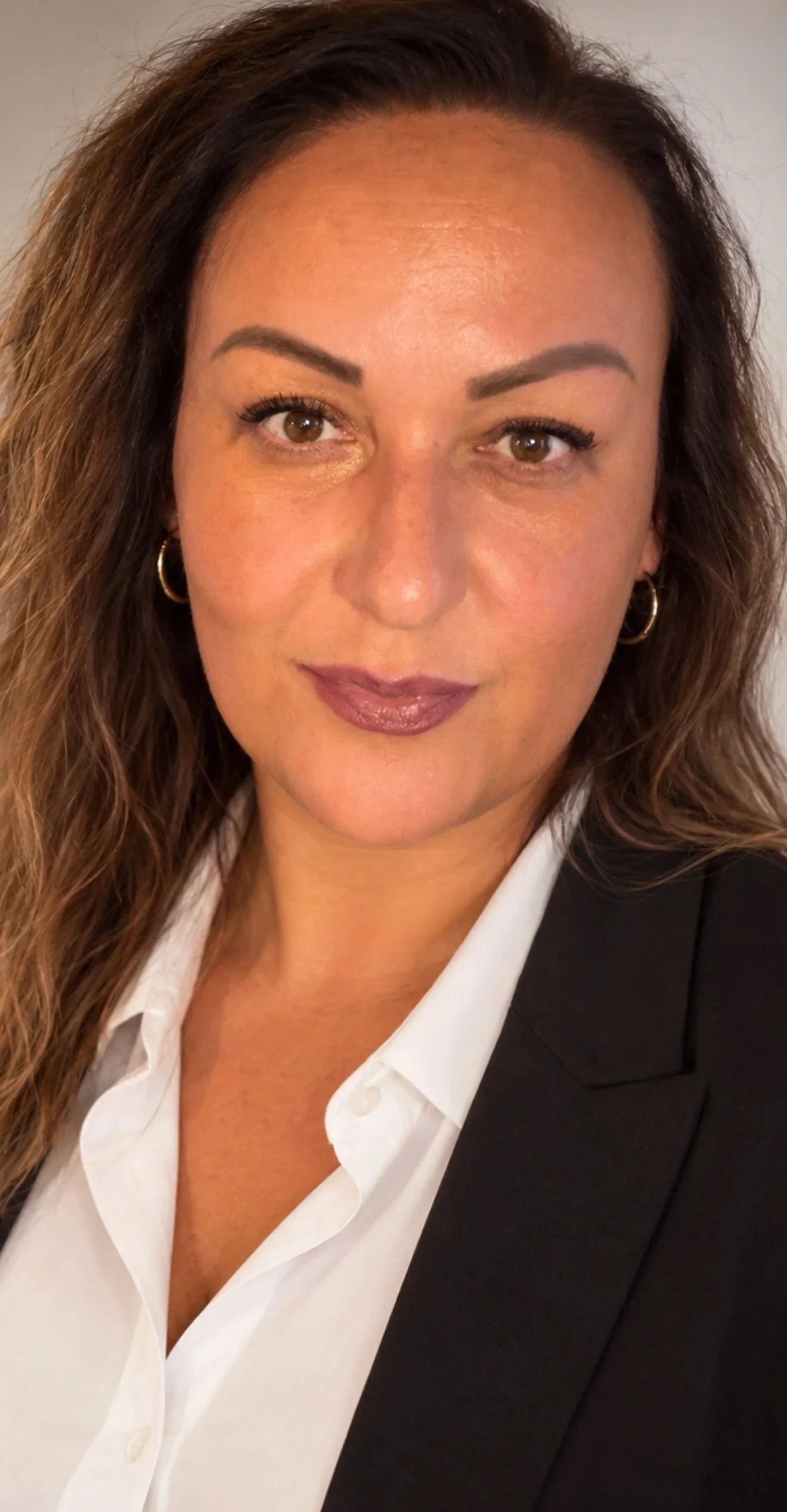 Porträt einer Frau mit braunen Haaren, braunen Augen, in Business-Kleidung, mit dunklem Blazer und weißem Hemd, lächelt leicht.