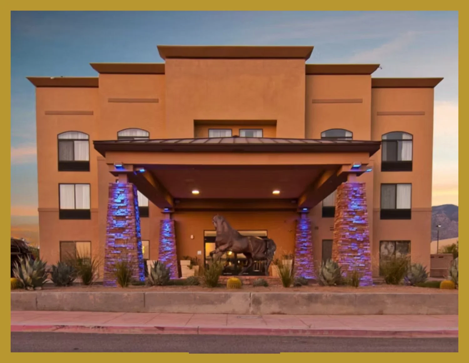 A multi-story building with a beige stucco exterior, arched windows, and a flat roof. The entrance features a covered porch supported by stone columns illuminated with blue lights. A large horse statue is situated in front of the building, surrounded by desert plants and cacti, with mountains and a sunset sky in the background.