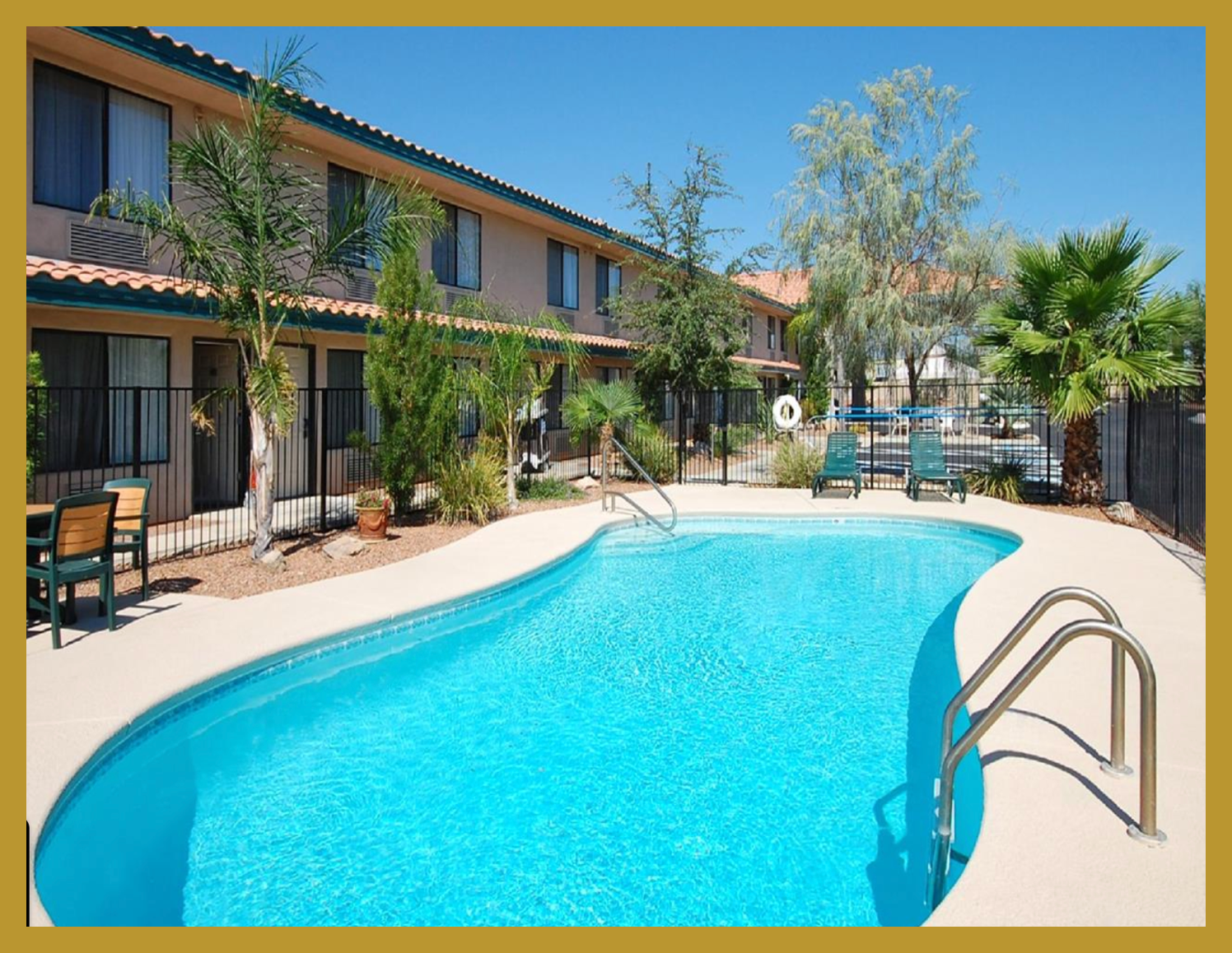 Apartment complex with a swimming pool surrounded by lounge chairs, palm trees, and a brown fence, under a clear blue sky.